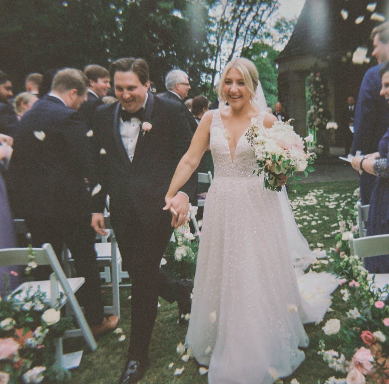 Joyful newlywed couple walking down the aisle as guests toss confetti at outdoor wedding.
