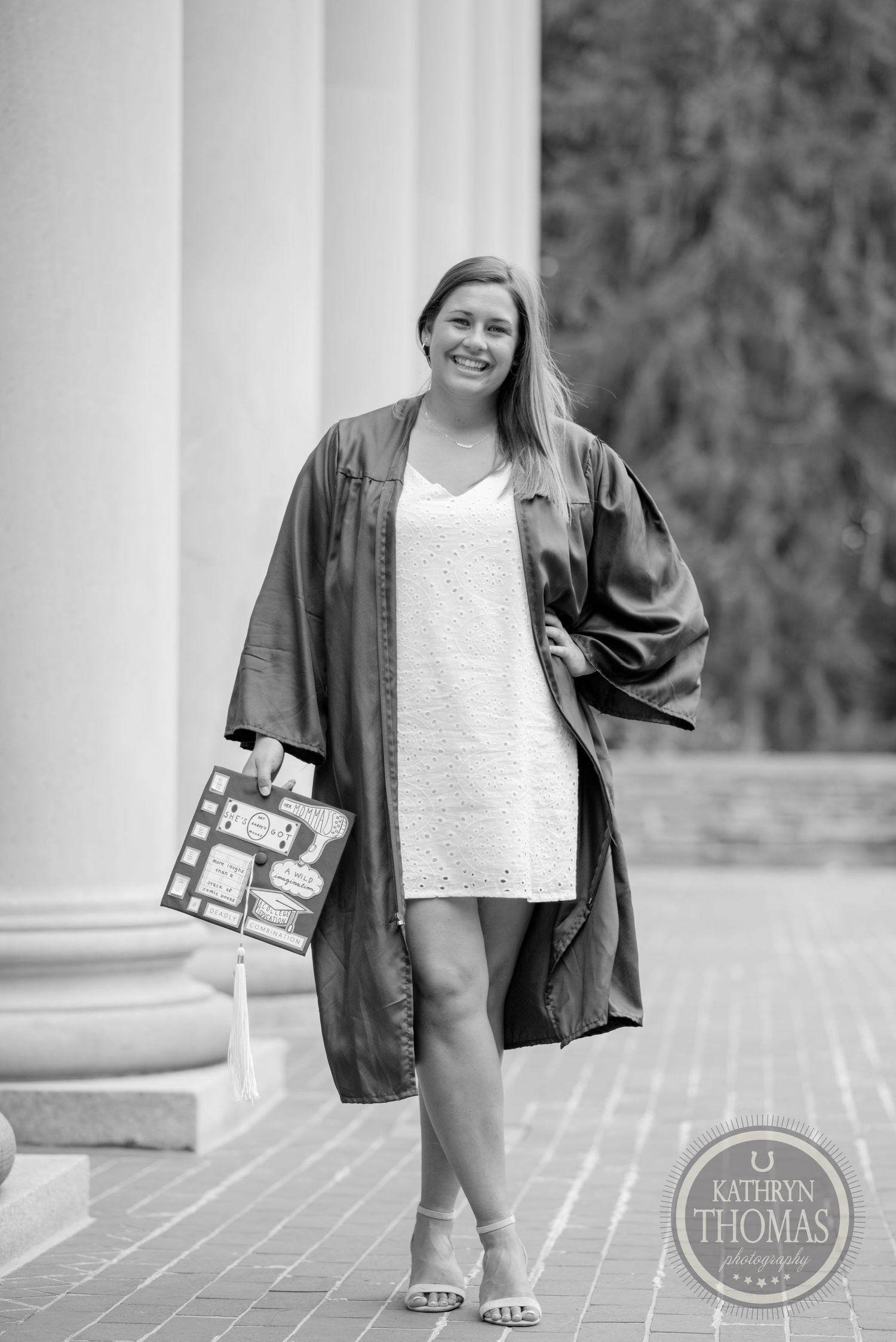 Black and white portrait of graduate in academic regalia holding diploma outside columned building.
