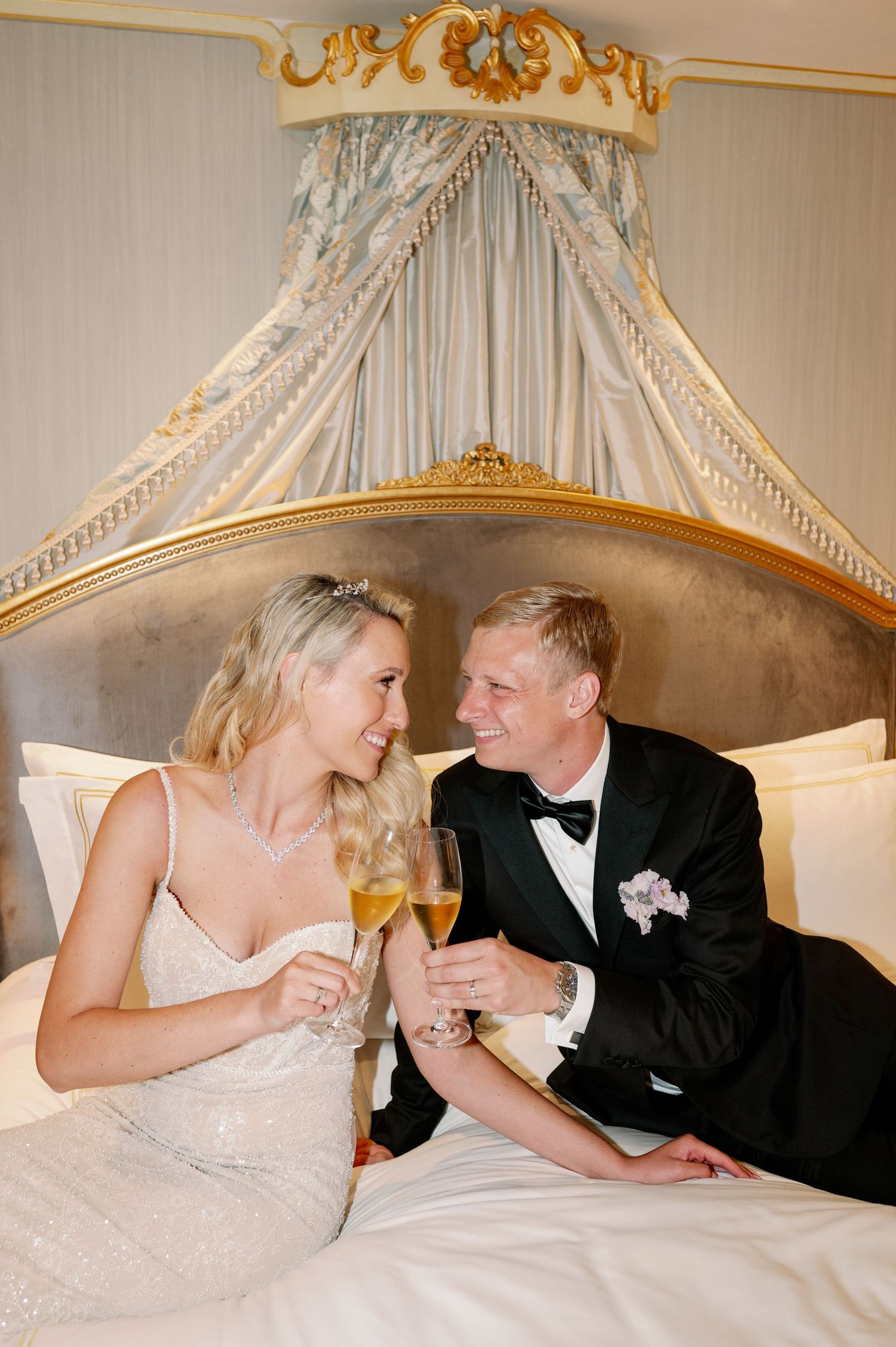Newlyweds share intimate moment under ornate canopy bed with champagne glasses.