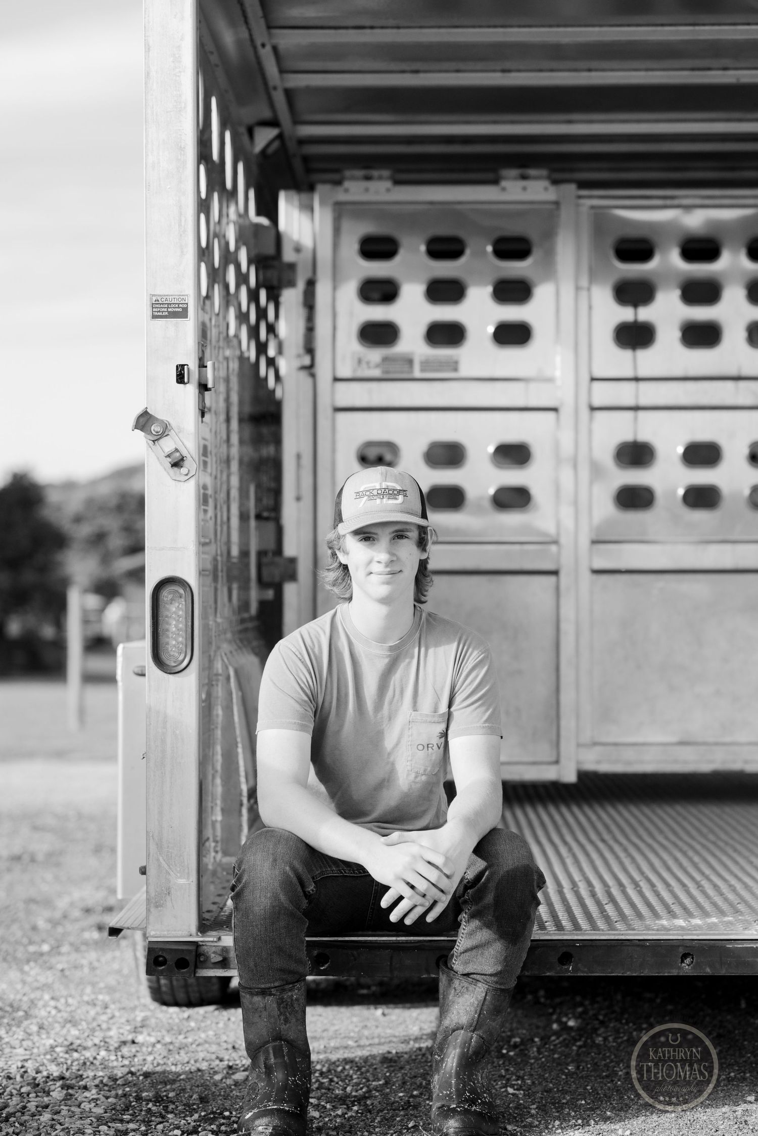 Black and white portrait of someone sitting on edge of livestock trailer wearing baseball cap.