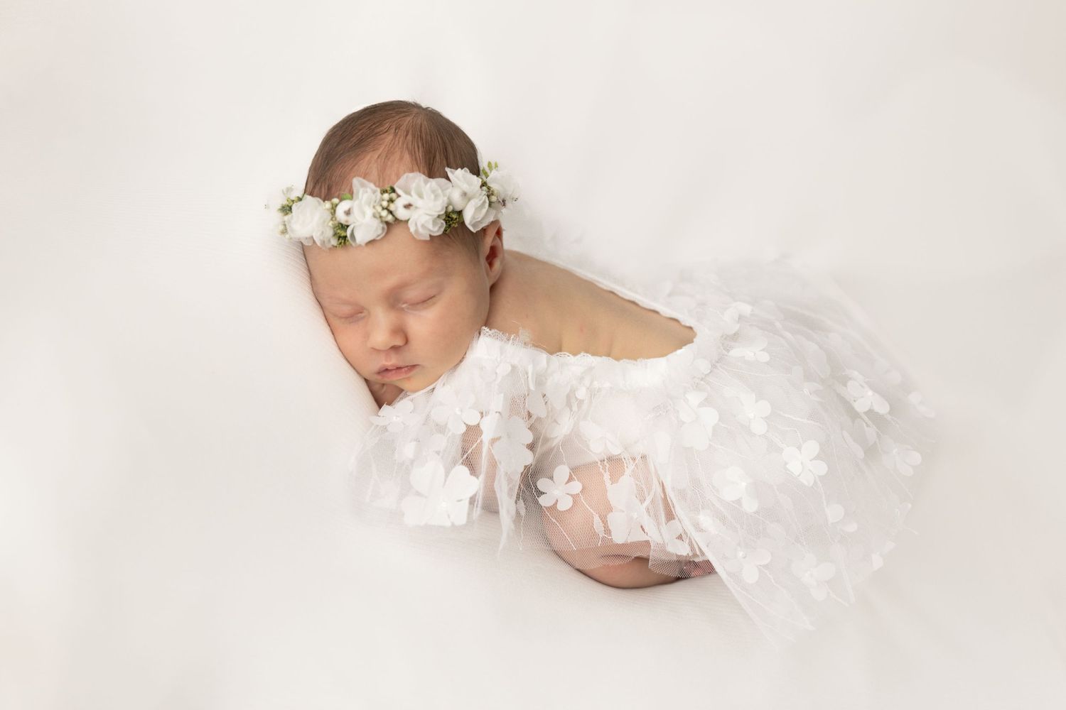 Newborn baby in delicate white wrap and flower crown sleeps peacefully on white background.