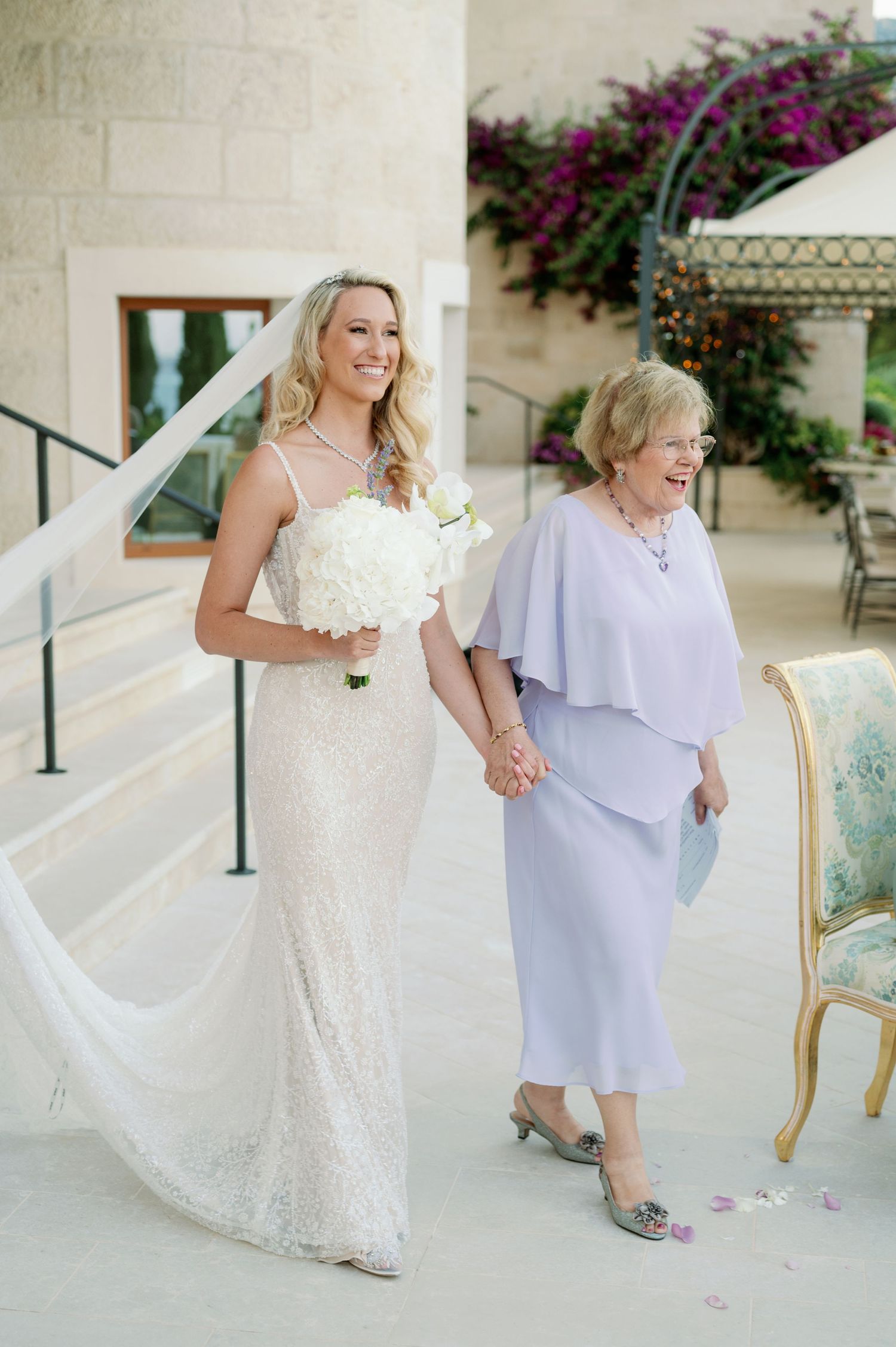 Elegant wedding scene on stone terrace with flowing veil and lavender dress.