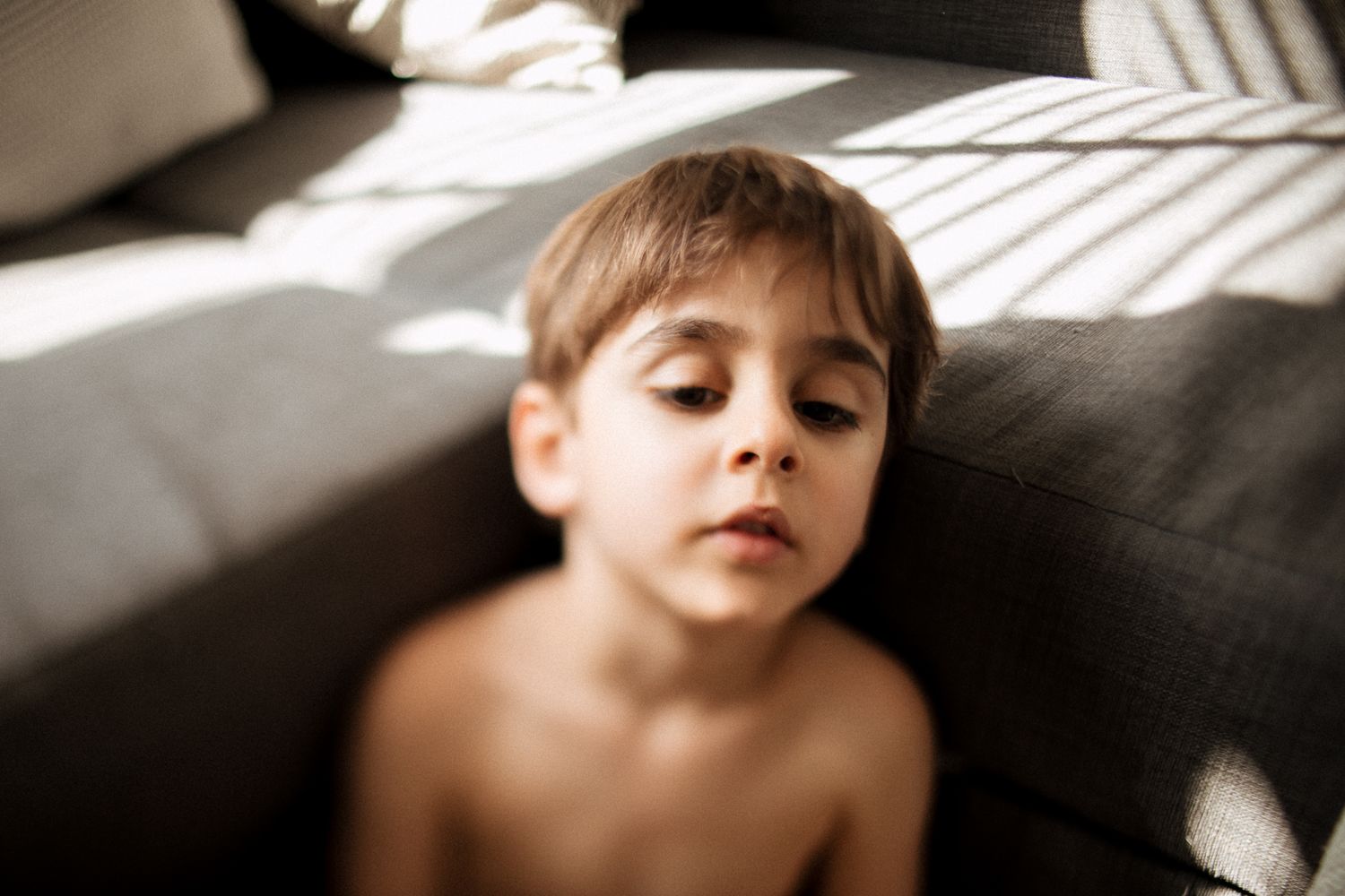 Young shirtless boy resting against a grey sofa in warm sunlight with eyes closed.