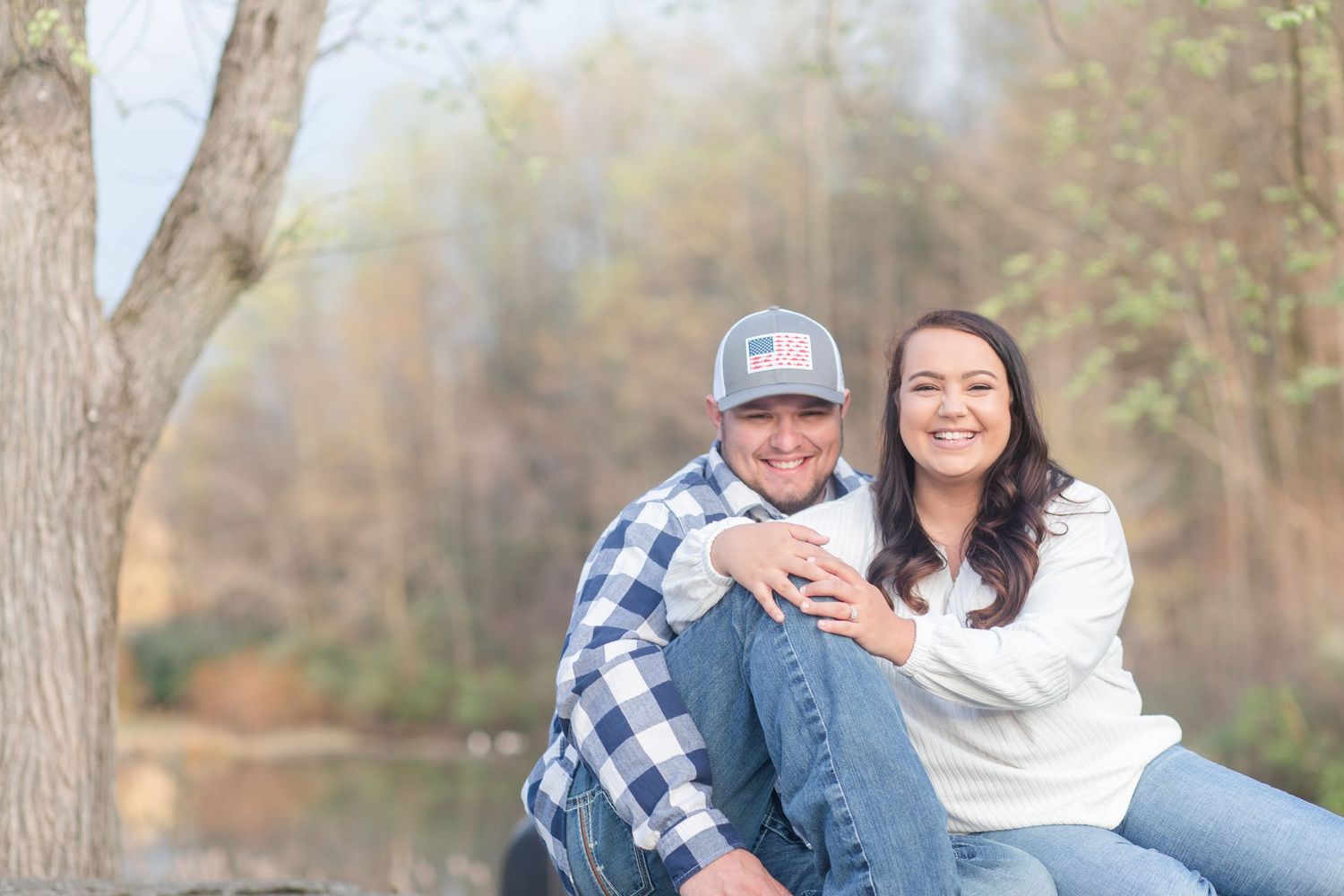 A couple sits together outdoors in casual attire with plaid shirt and white sweater near a tree.