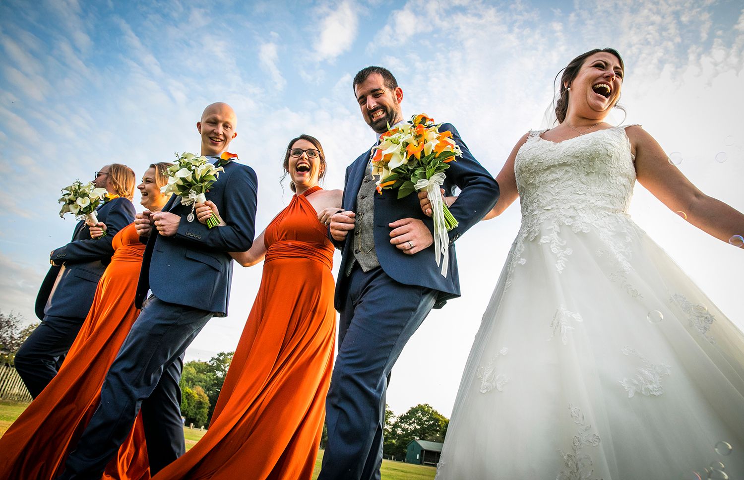 Wedding party wearing navy and orange attire pose together outdoors with bright sunlight and joyful expressions.