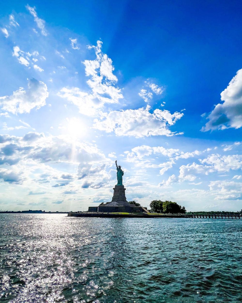 Statue of Liberty stands against bright blue sky with sunburst and scattered clouds.