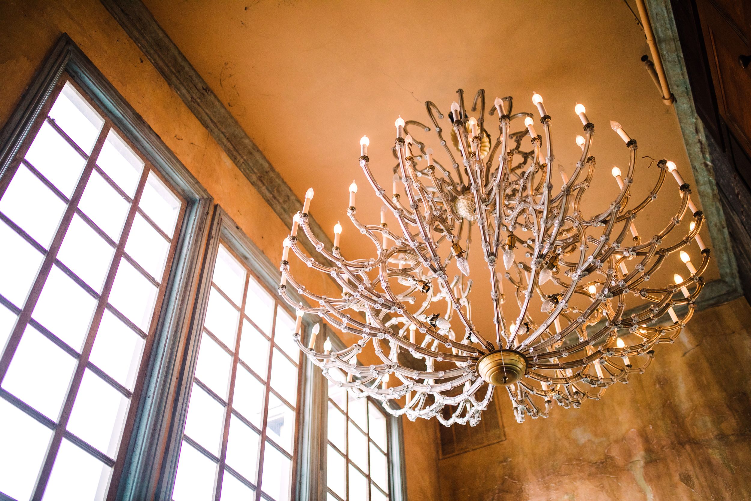 Elegant branch-style chandelier with candle lights hanging from an amber-colored ceiling near tall windows.