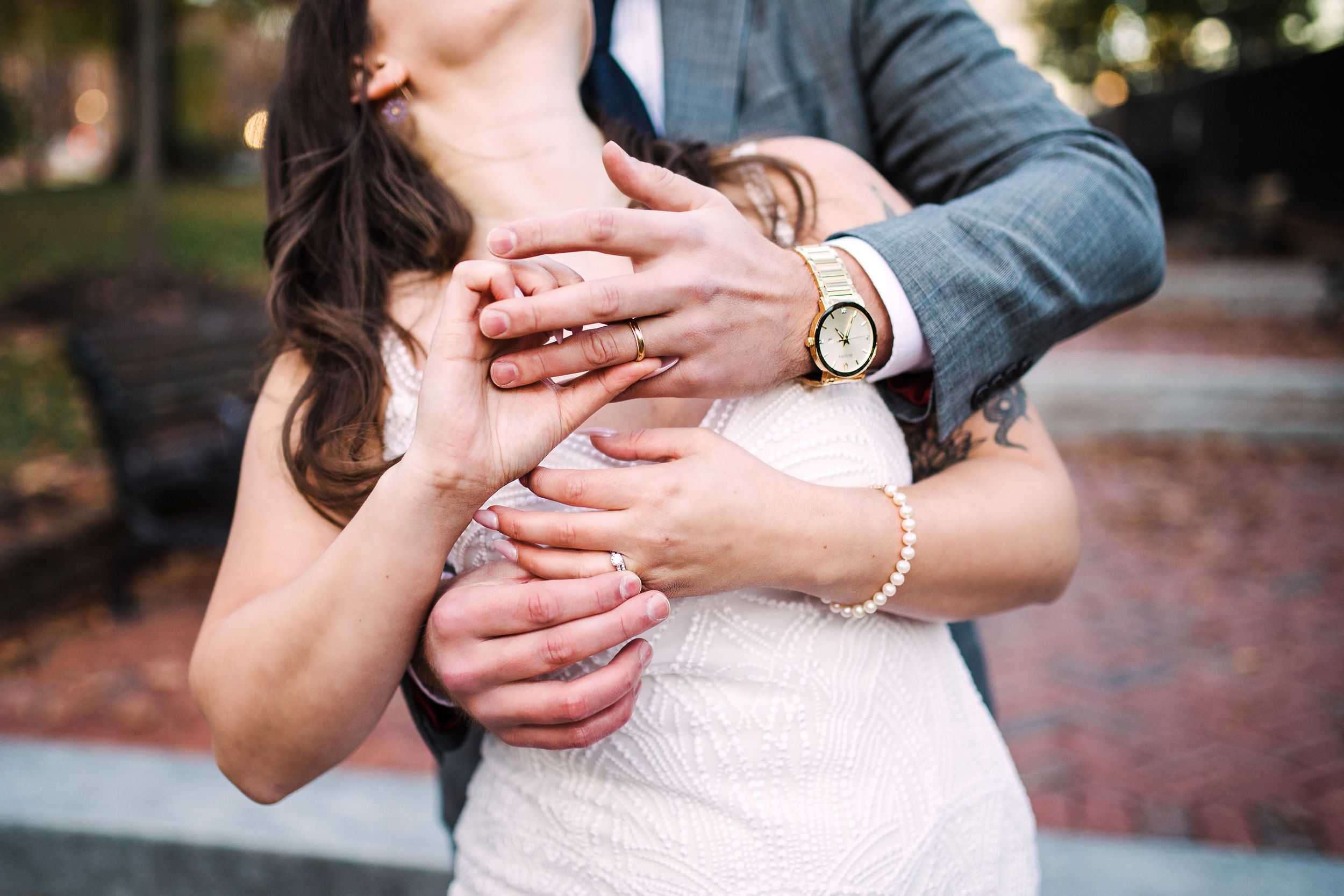 Groom embracing bride from behind showing wedding rings and gold watch in romantic close-up shot.
