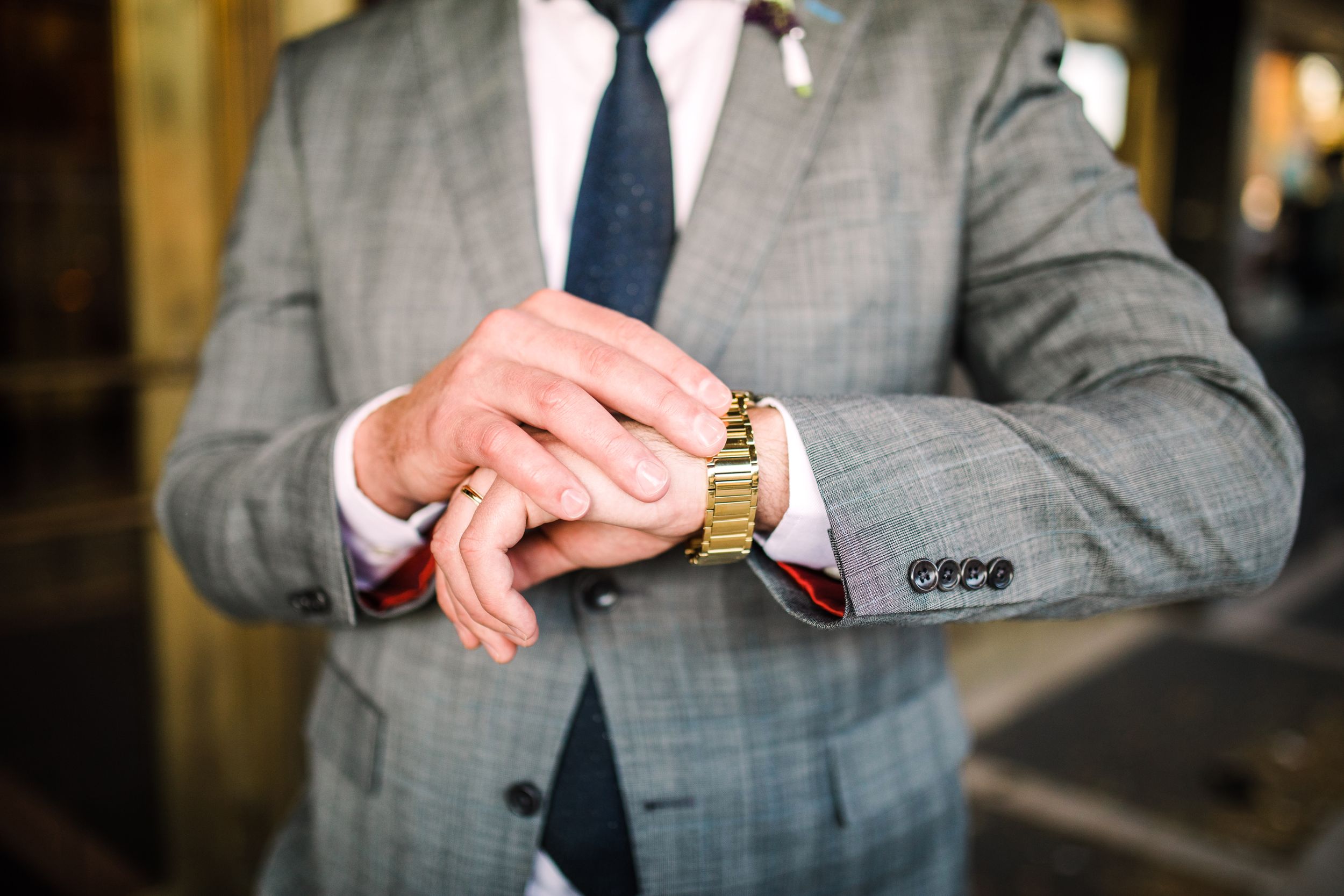 Groom in gray plaid suit adjusting gold watch with navy tie in close-up detail shot.