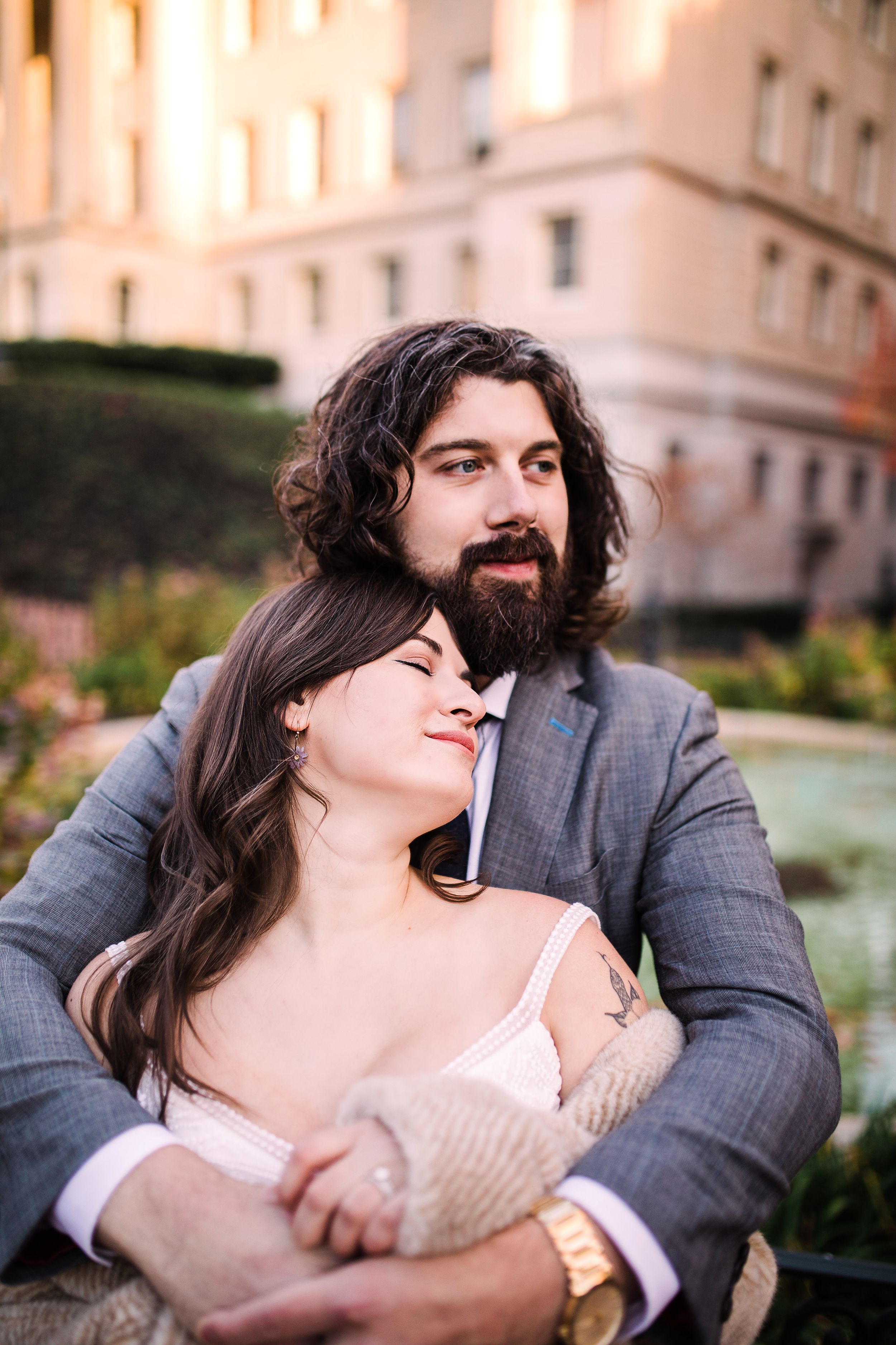 Bearded groom holding bride close while sitting outdoors near fountain at golden hour.