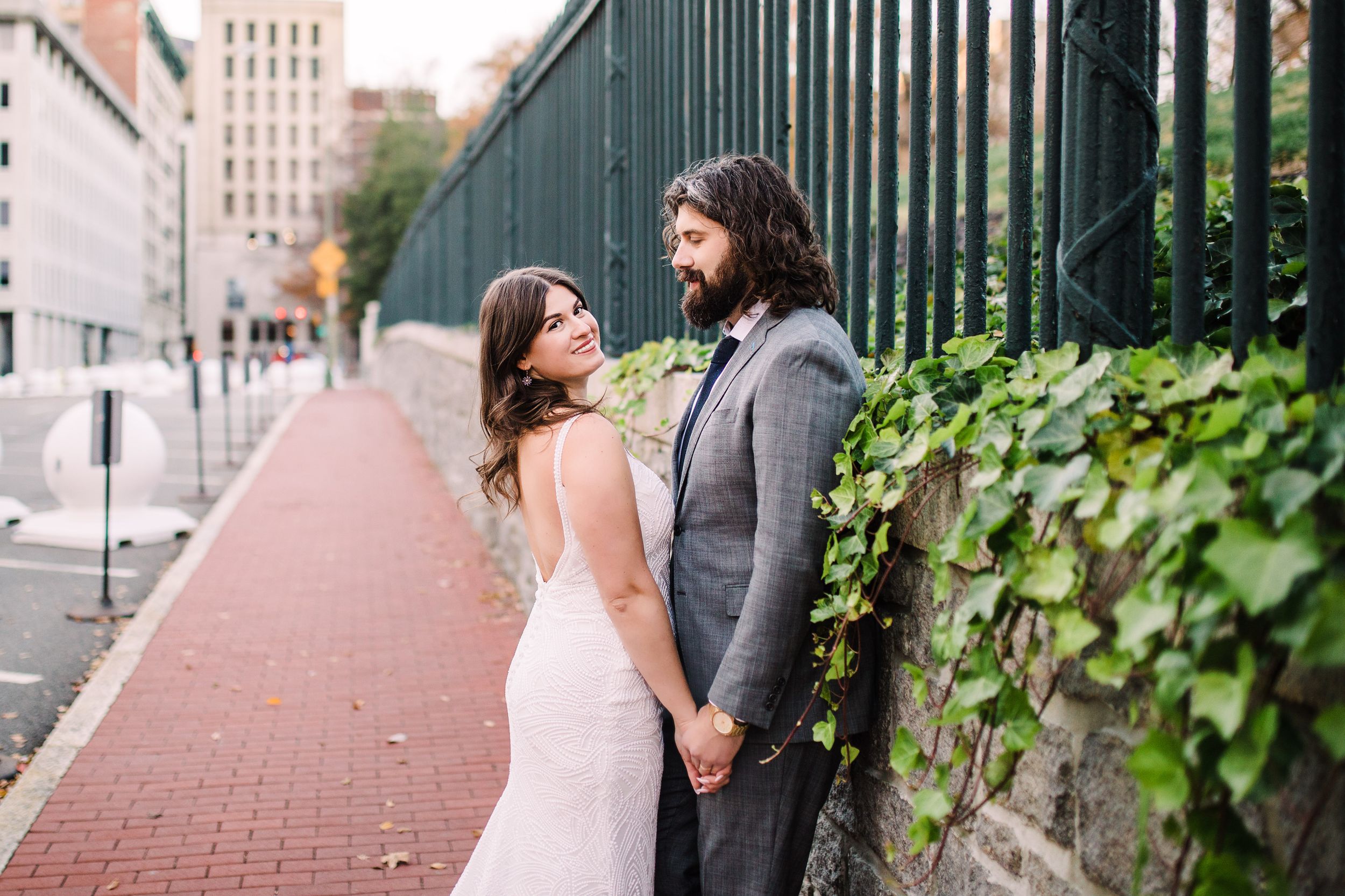 Bride and groom holding hands by ivy-covered stone wall on city sidewalk.