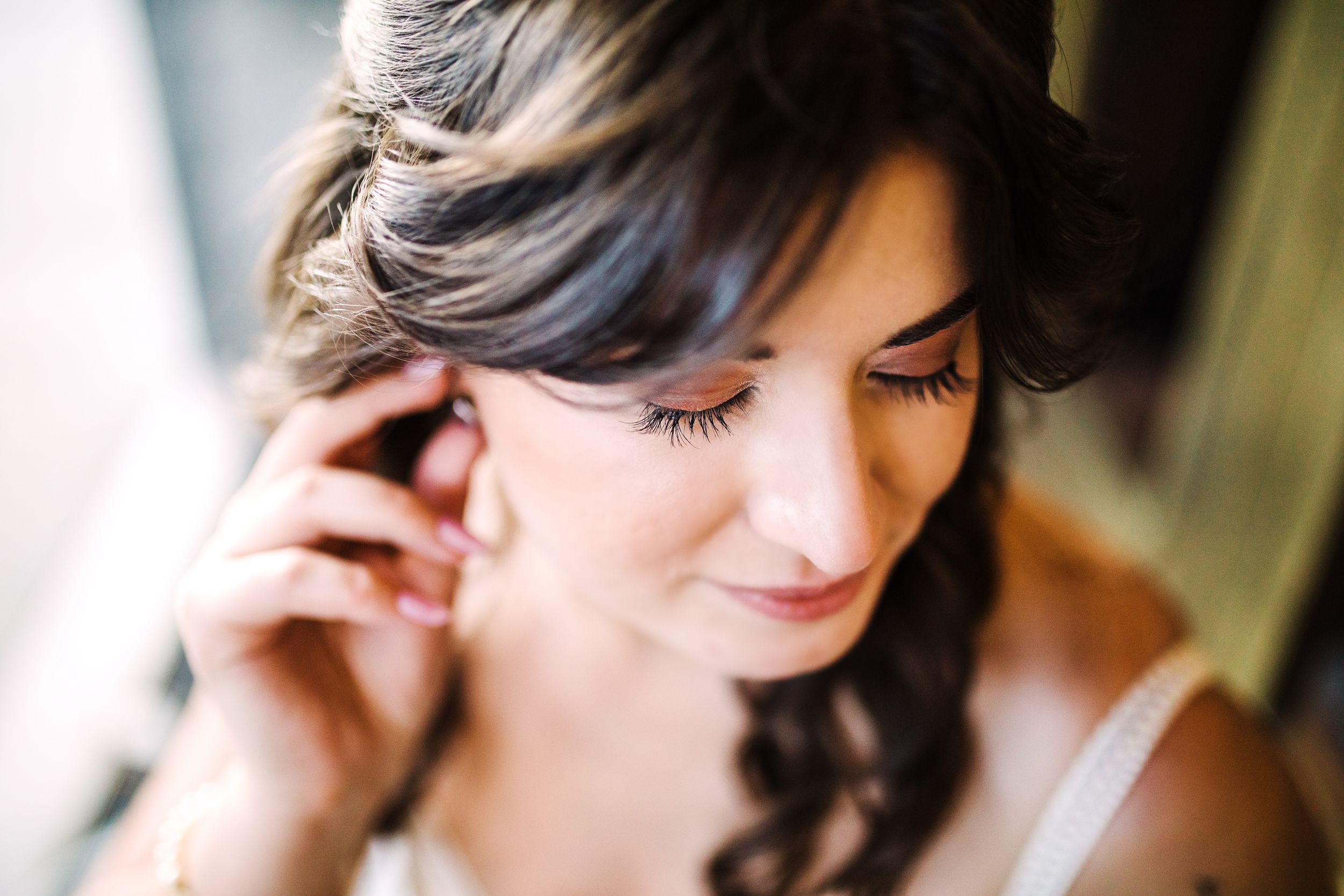 Bride putting on earring during wedding day preparations, soft natural light.