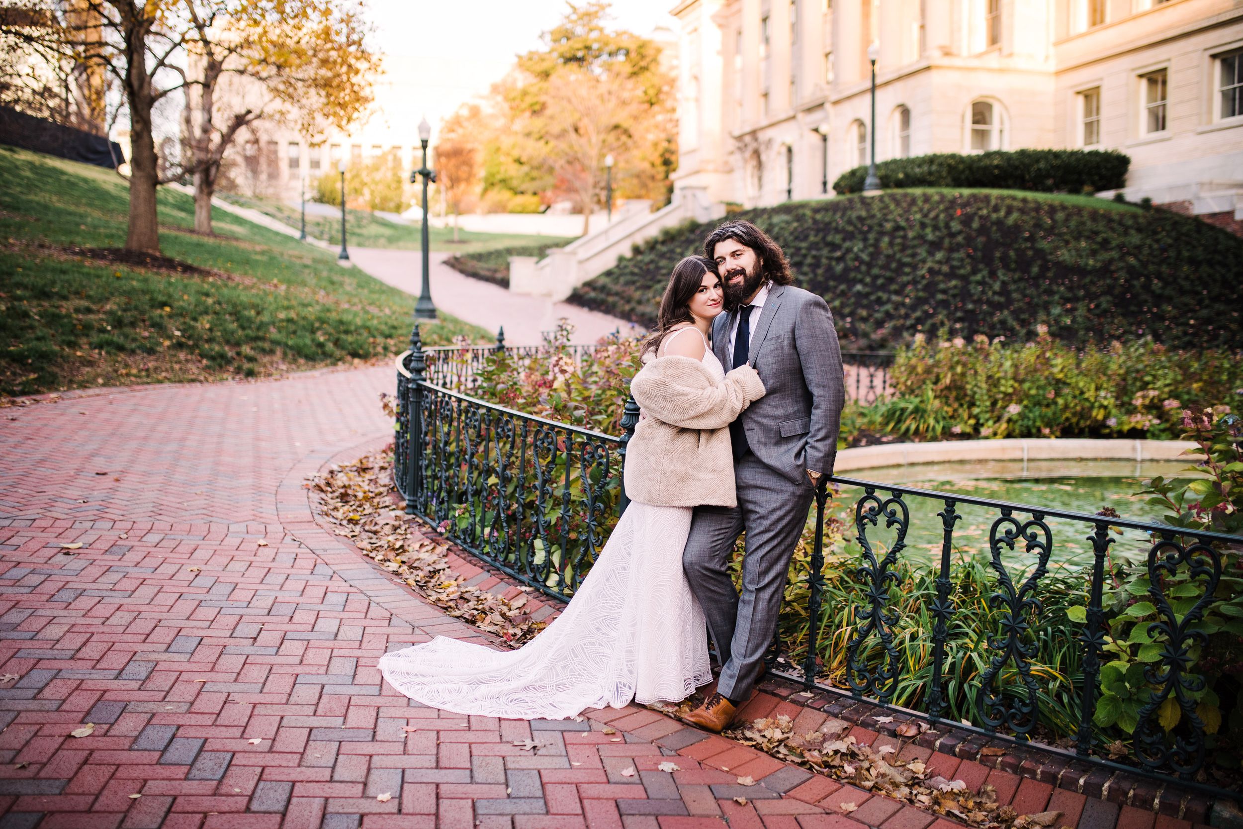 Bride and groom posing by ornate iron fence with autumn trees and historic building backdrop.