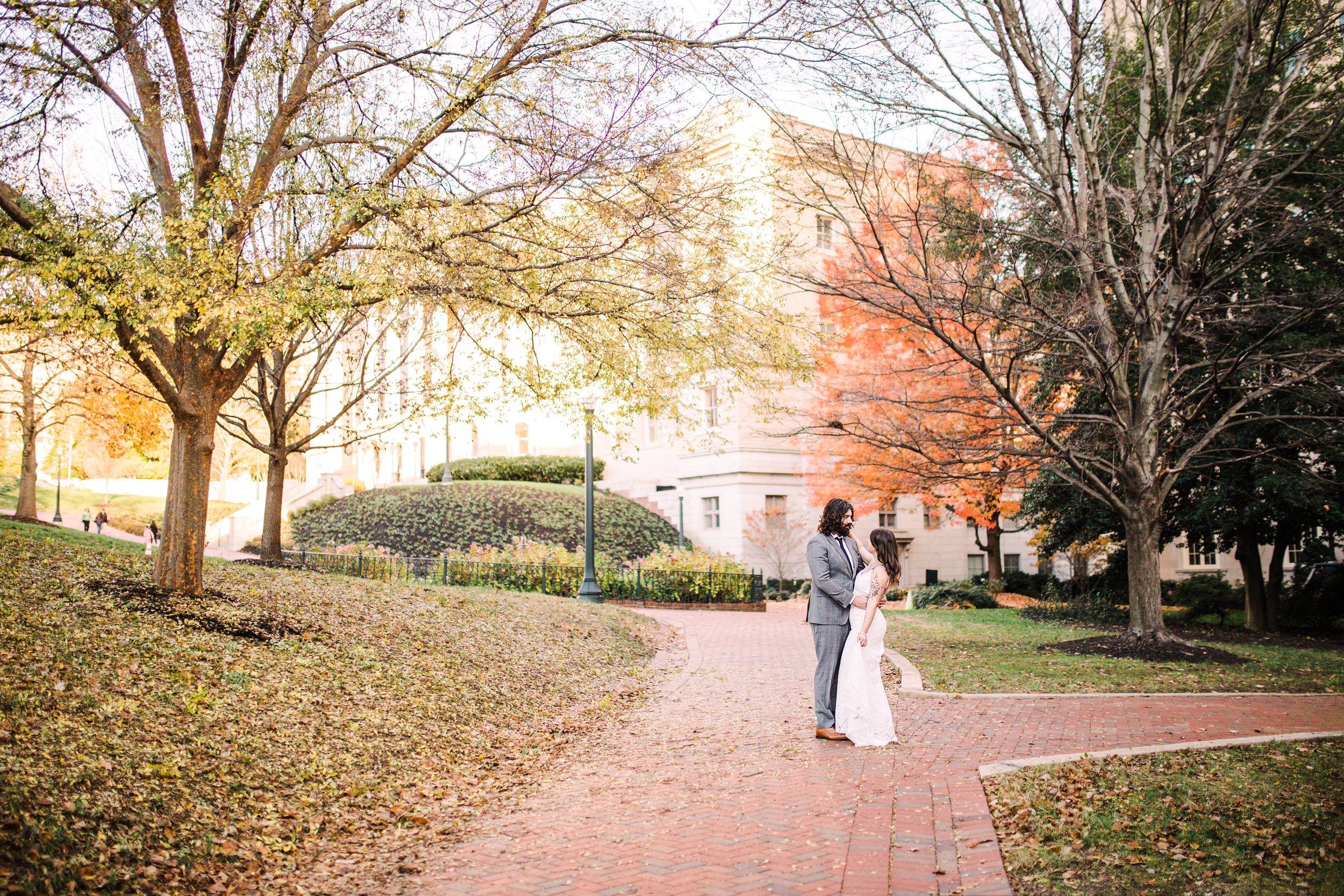 Bride and groom walking hand in hand on brick path surrounded by colorful fall foliage.