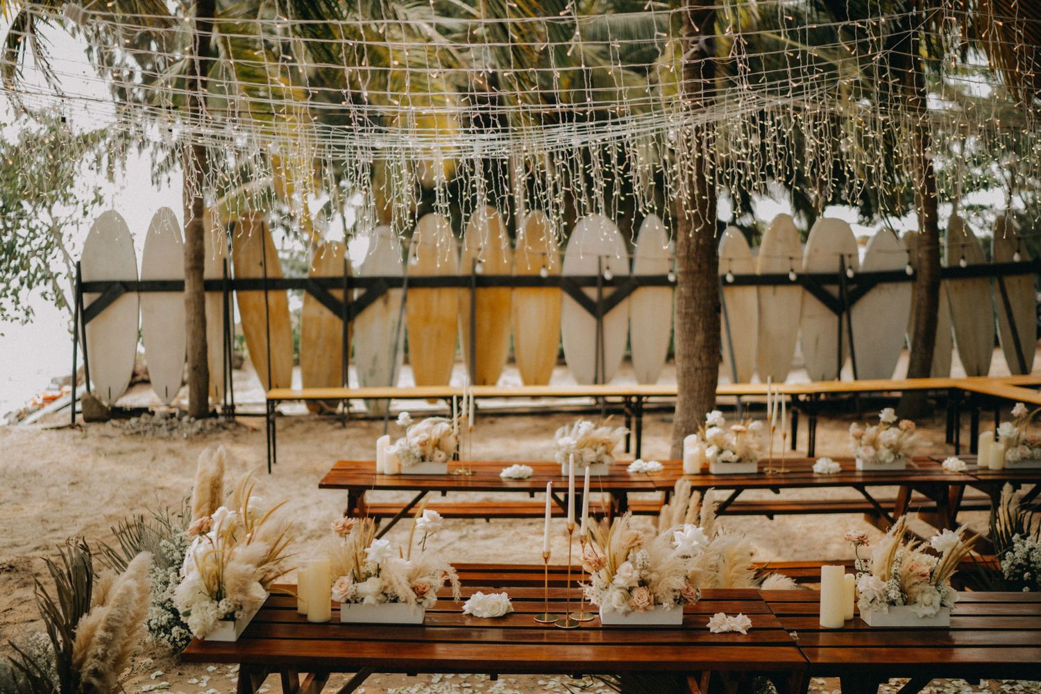 Rustic outdoor wedding reception with wooden tables, pampas grass, and surfboards backdrop.