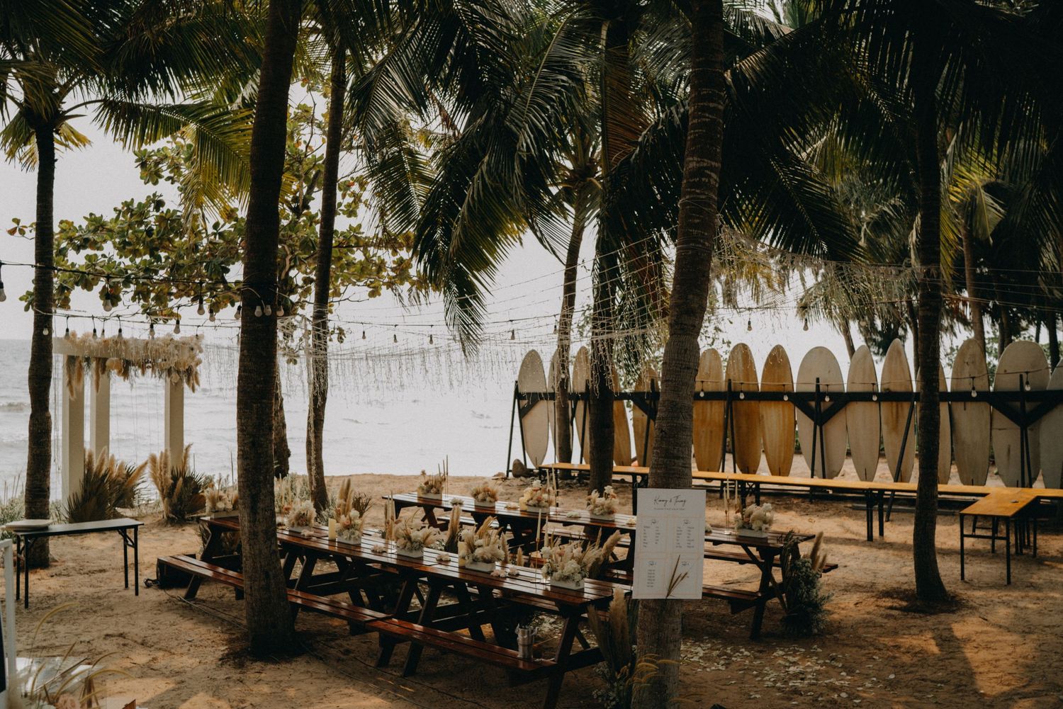 Tropical beach wedding reception with wooden tables set among palm trees and surfboards.