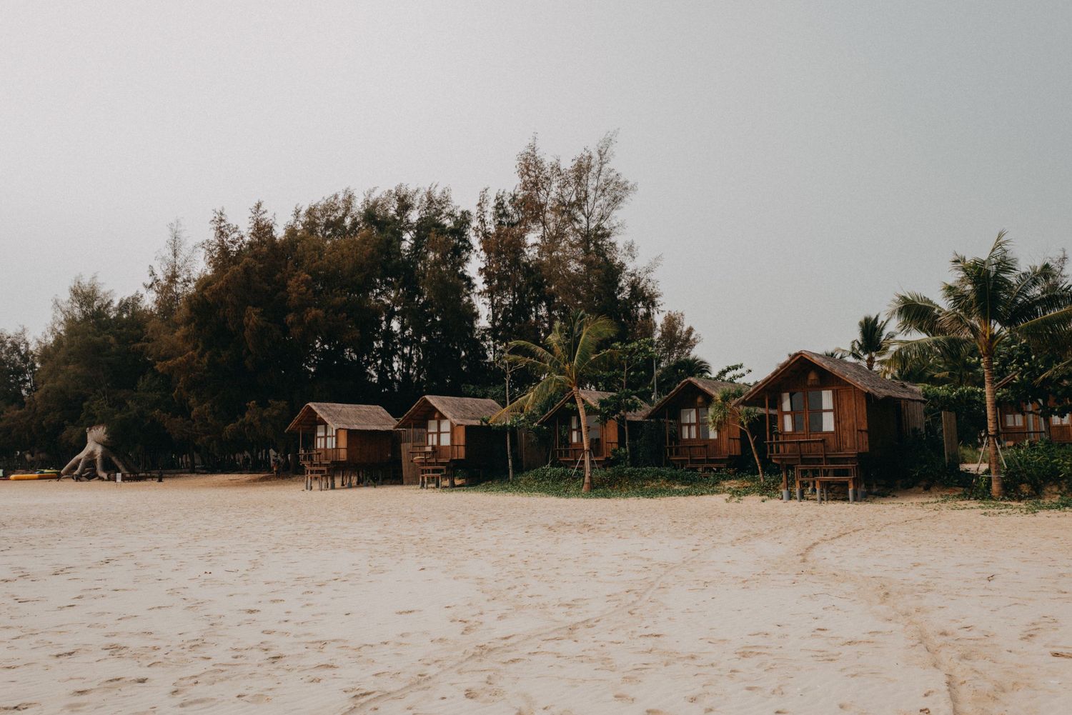 Rustic wooden beach bungalows on white sandy shore surrounded by trees.
