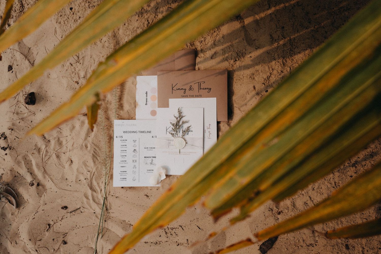 Wedding stationery flatlay on sand framed by tropical palm leaves.