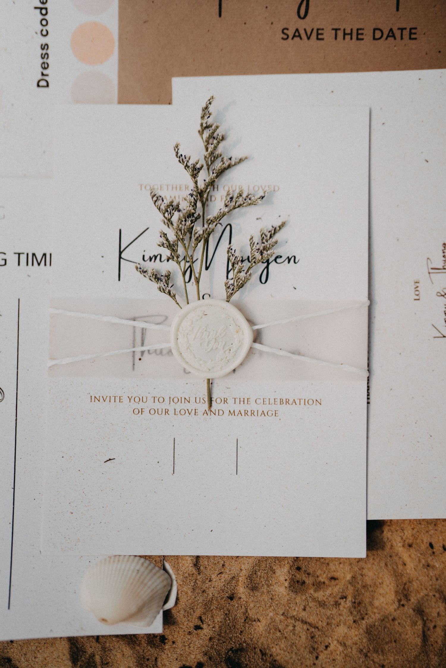 Elegant wedding invitation with wax seal, ribbon, and dried botanicals on sandy beach surface.