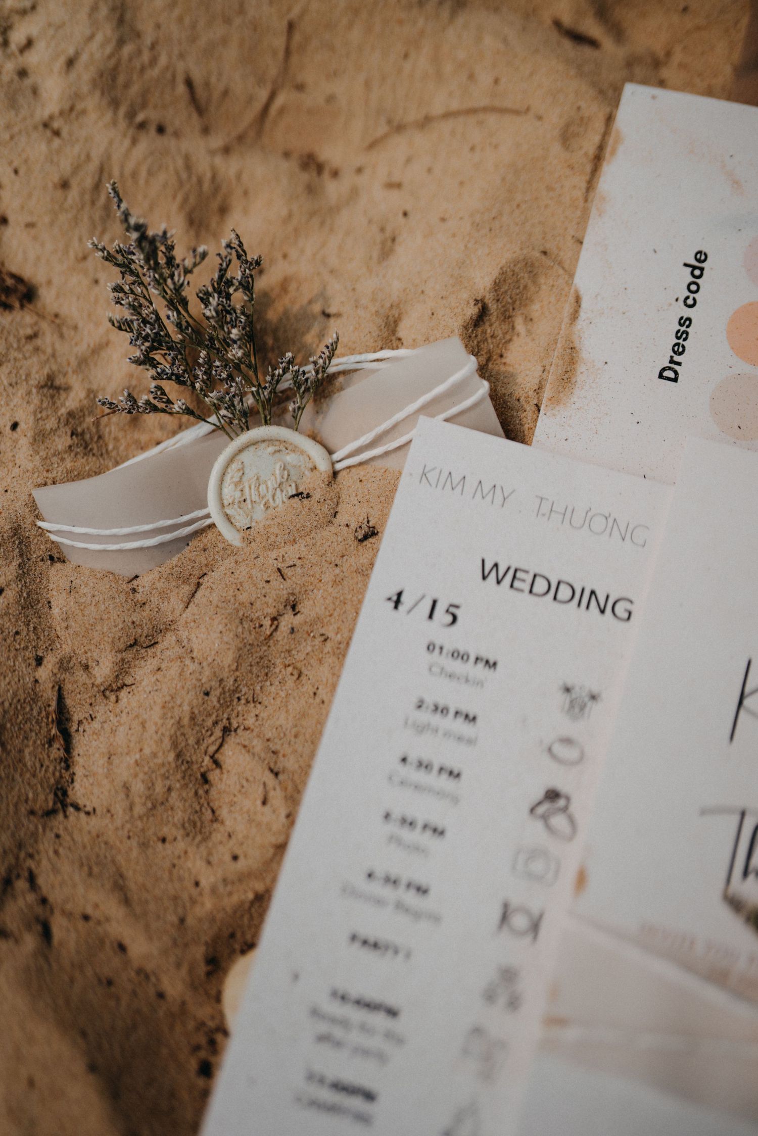Wedding timeline card for Kimmy and Thuong on 4/15 with wax seal and dried herbs on sand.