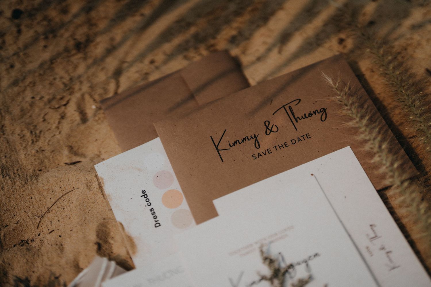Kraft envelope save the date card for Kimmy and Thuong displayed on sandy beach surface.