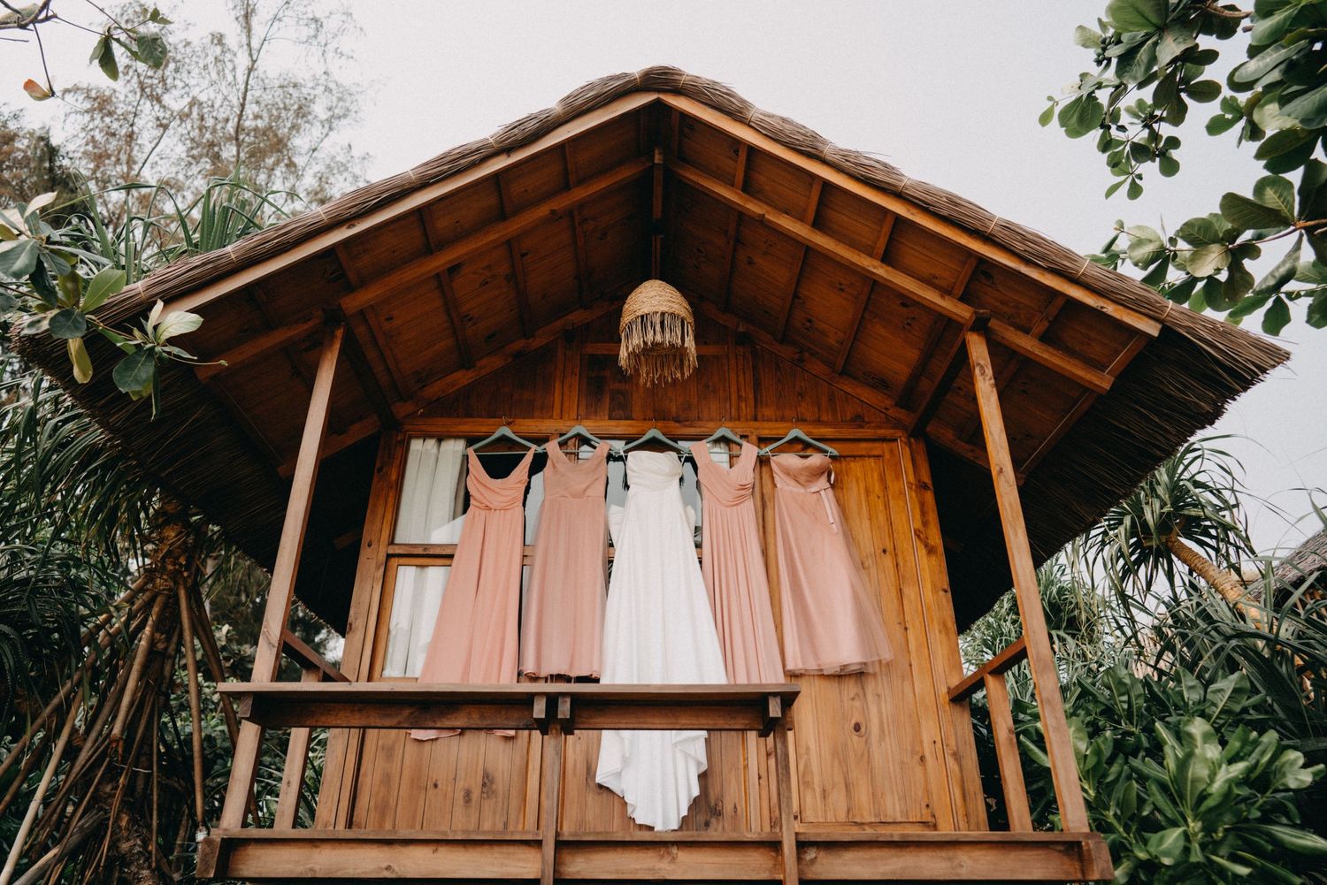 White wedding dress and pink bridesmaid gowns hanging on rustic wooden tropical beach bungalow.