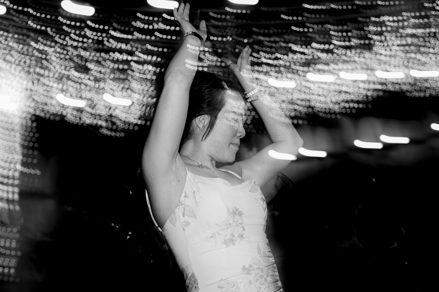 Black and white close-up of a woman dancing with arms raised under glowing string lights at a wedding.