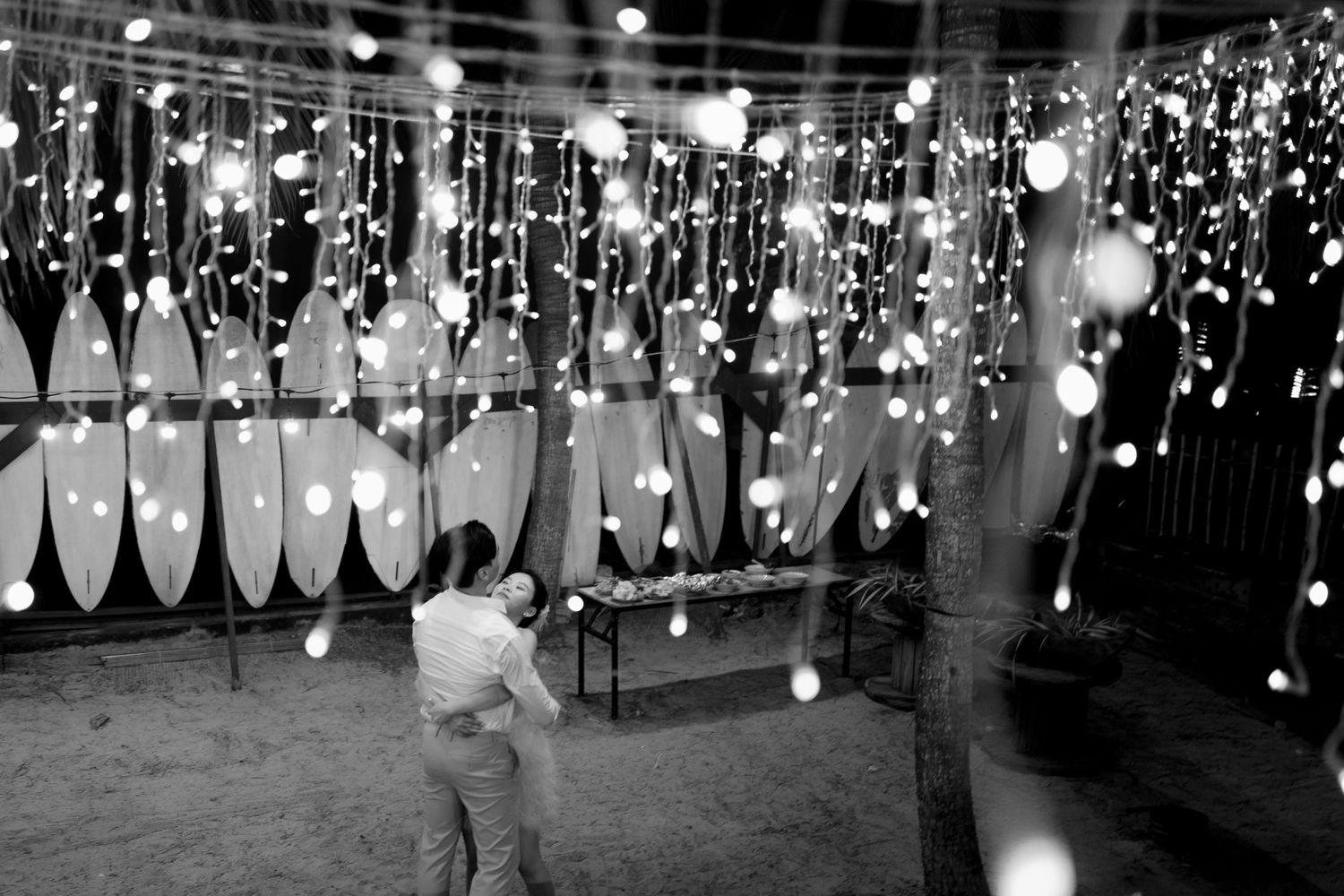 Black and white photo of a couple embracing under cascading string lights and lanterns at an outdoor wedding.