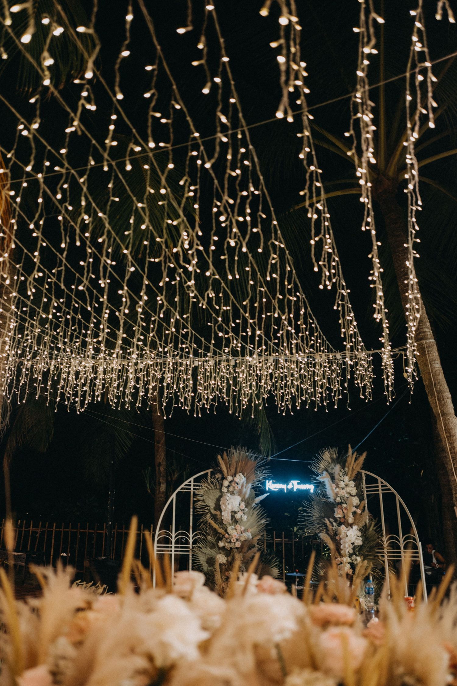 Stunning wedding arch with neon sign under cascading fairy lights surrounded by pampas grass floral arrangements.
