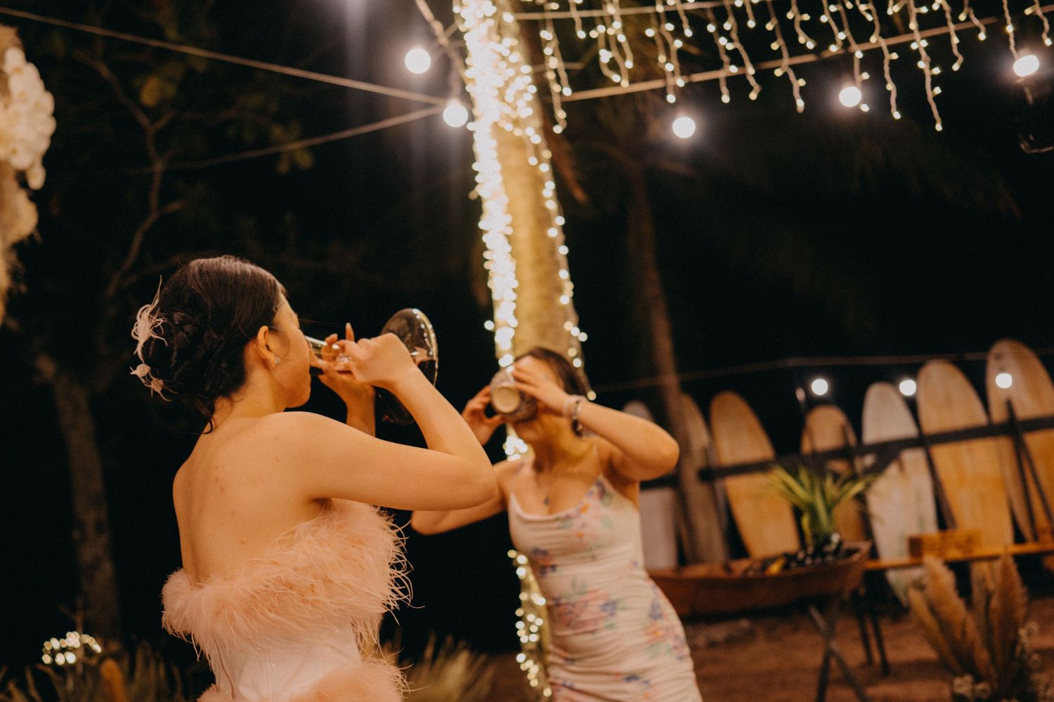 Two brides in white dresses toasting champagne under string lights at an outdoor wedding celebration.