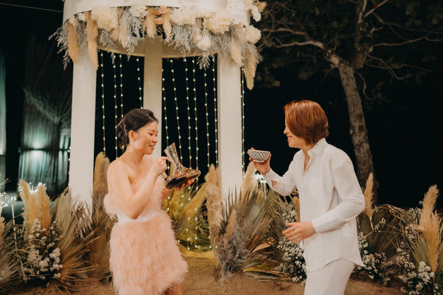 Two brides in pink feather and white suits toasting champagne under a floral arch at their wedding.