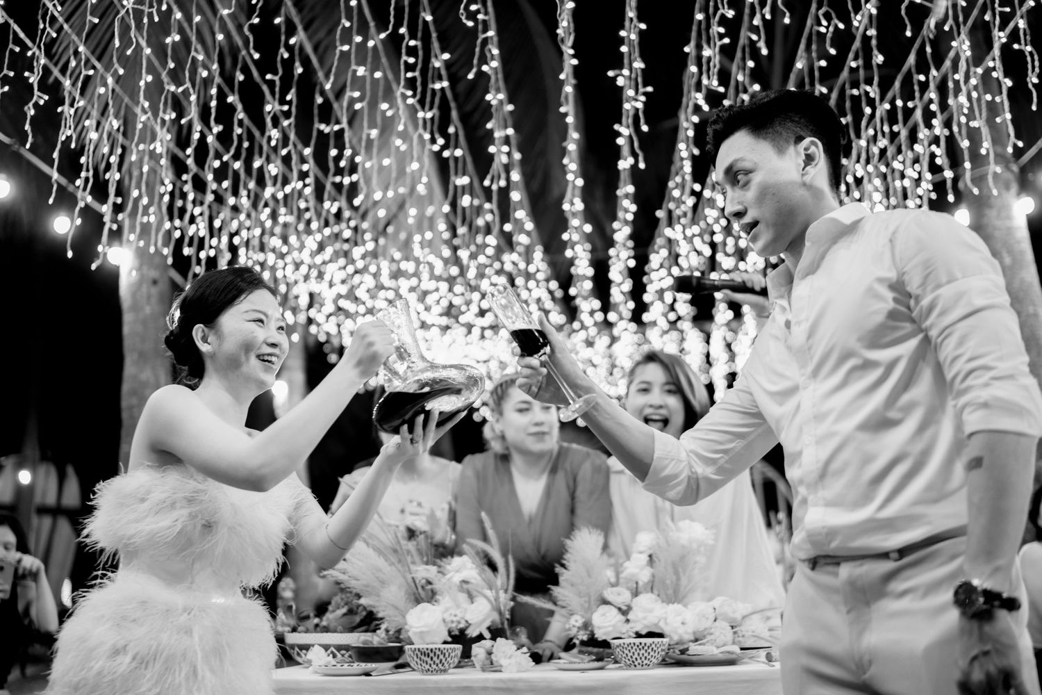 Black and white photo of brides and guests joyfully toasting at a wedding reception under fairy lights.