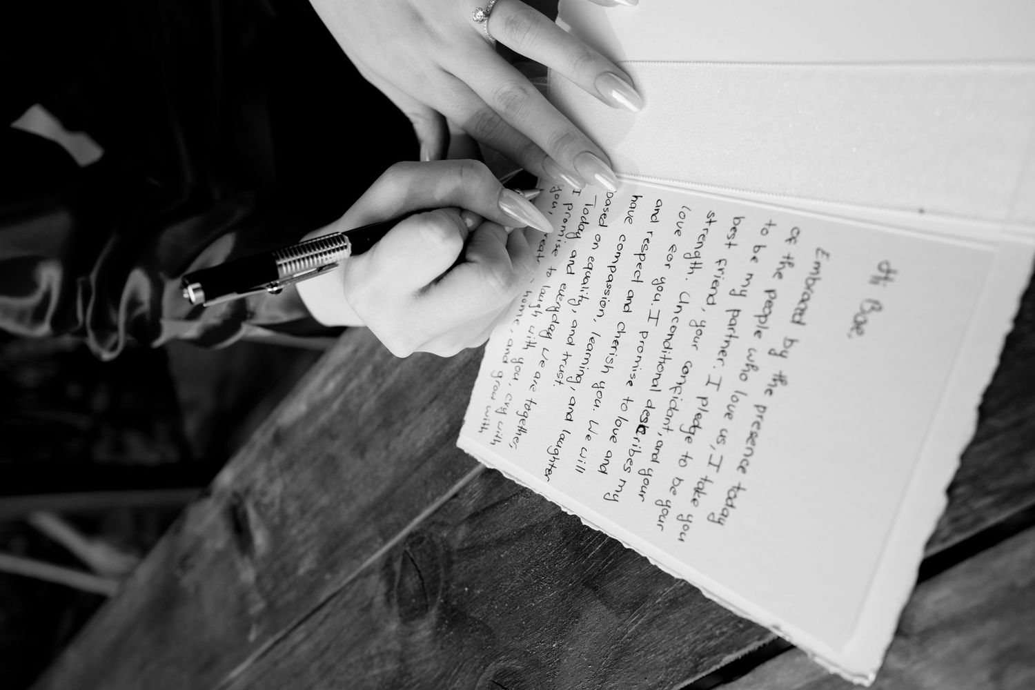 Black and white close-up of hands writing heartfelt wedding vows with pen on paper.