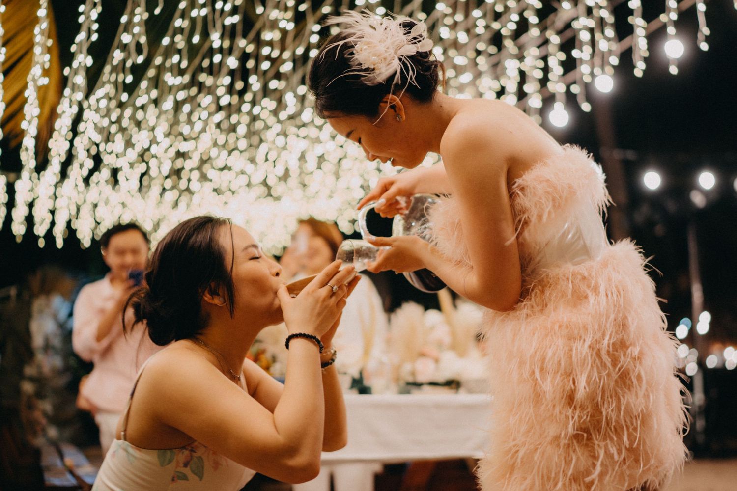 Bride in pink feather dress feeding wedding cake to a guest under glowing string lights at reception.