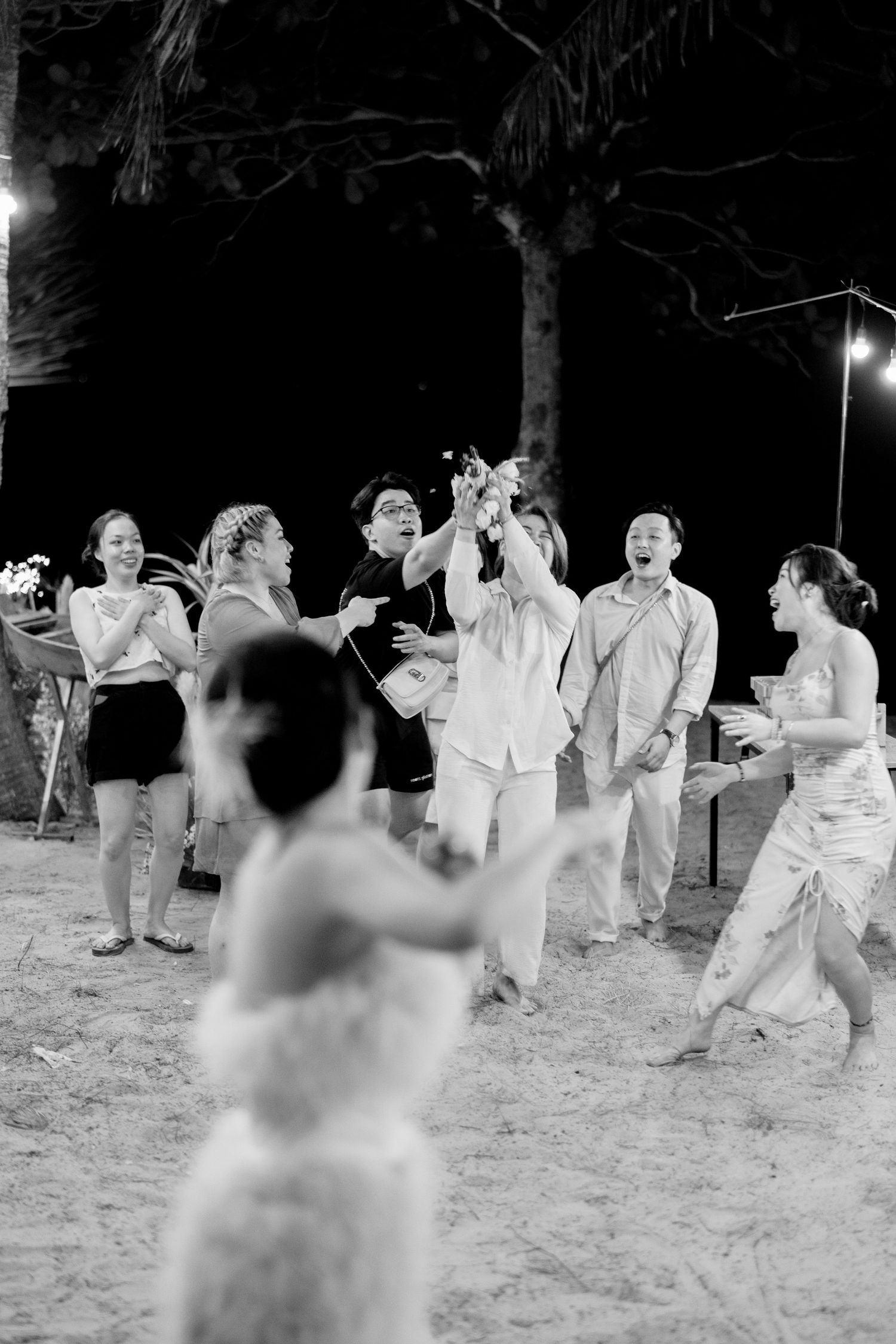 Black and white photo of wedding guests cheering during bouquet toss on sandy outdoor venue at night.