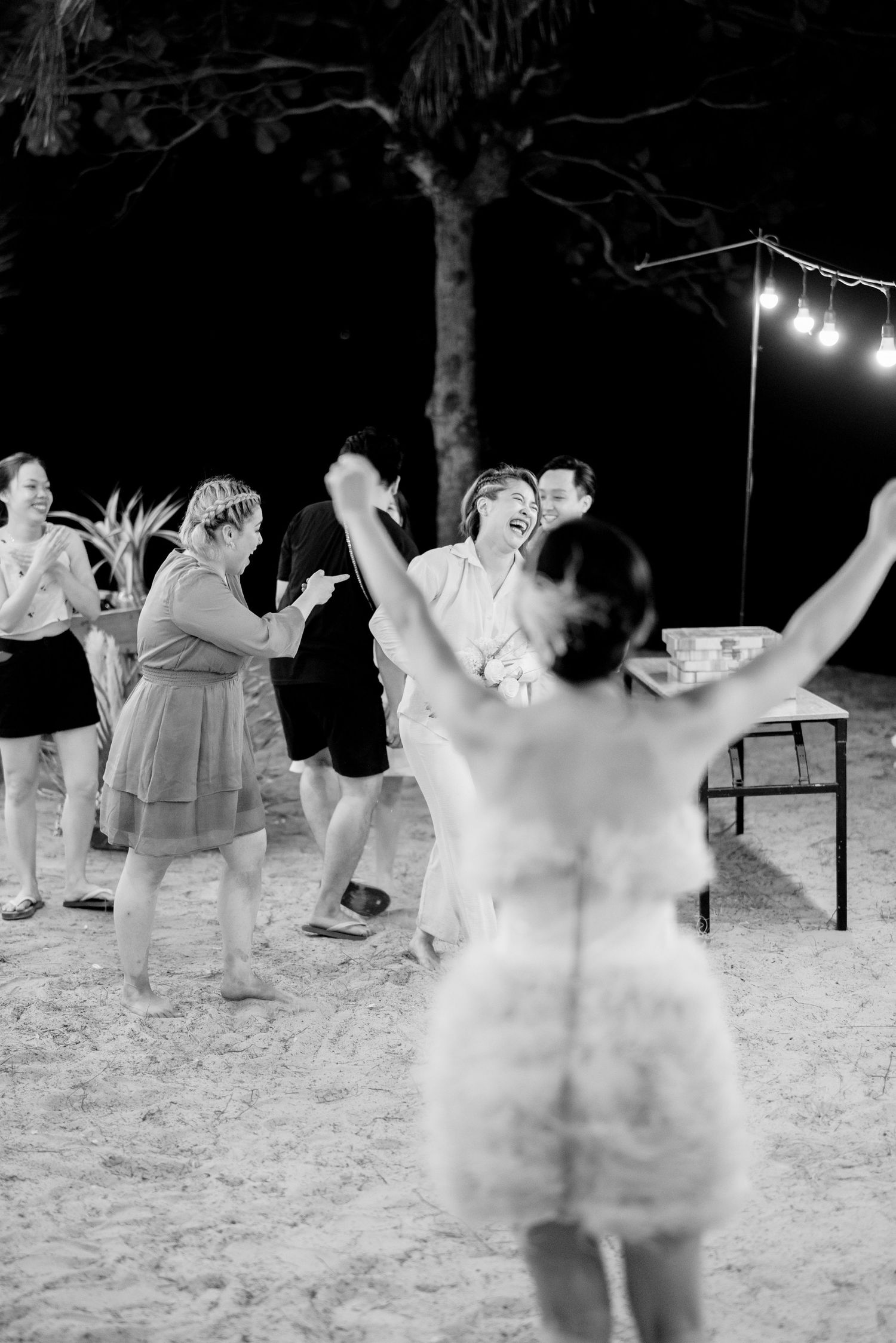 Guests laugh and dance joyfully on sandy beach at night under string lights during outdoor wedding reception.