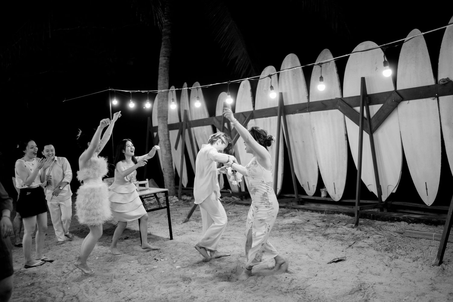 Wedding guests dance energetically on beach at night with surfboards displayed against wall in background.