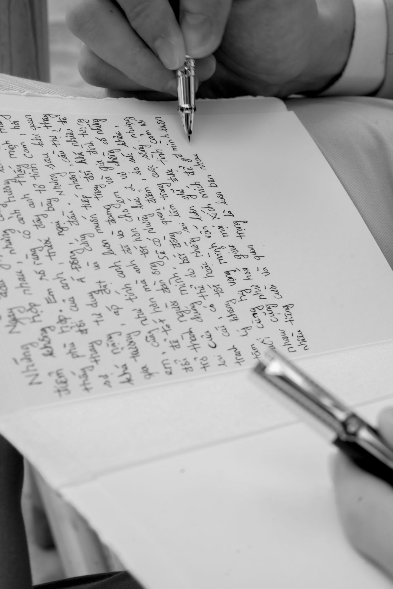 Black and white close-up of groom writing handwritten wedding vows with fountain pen.