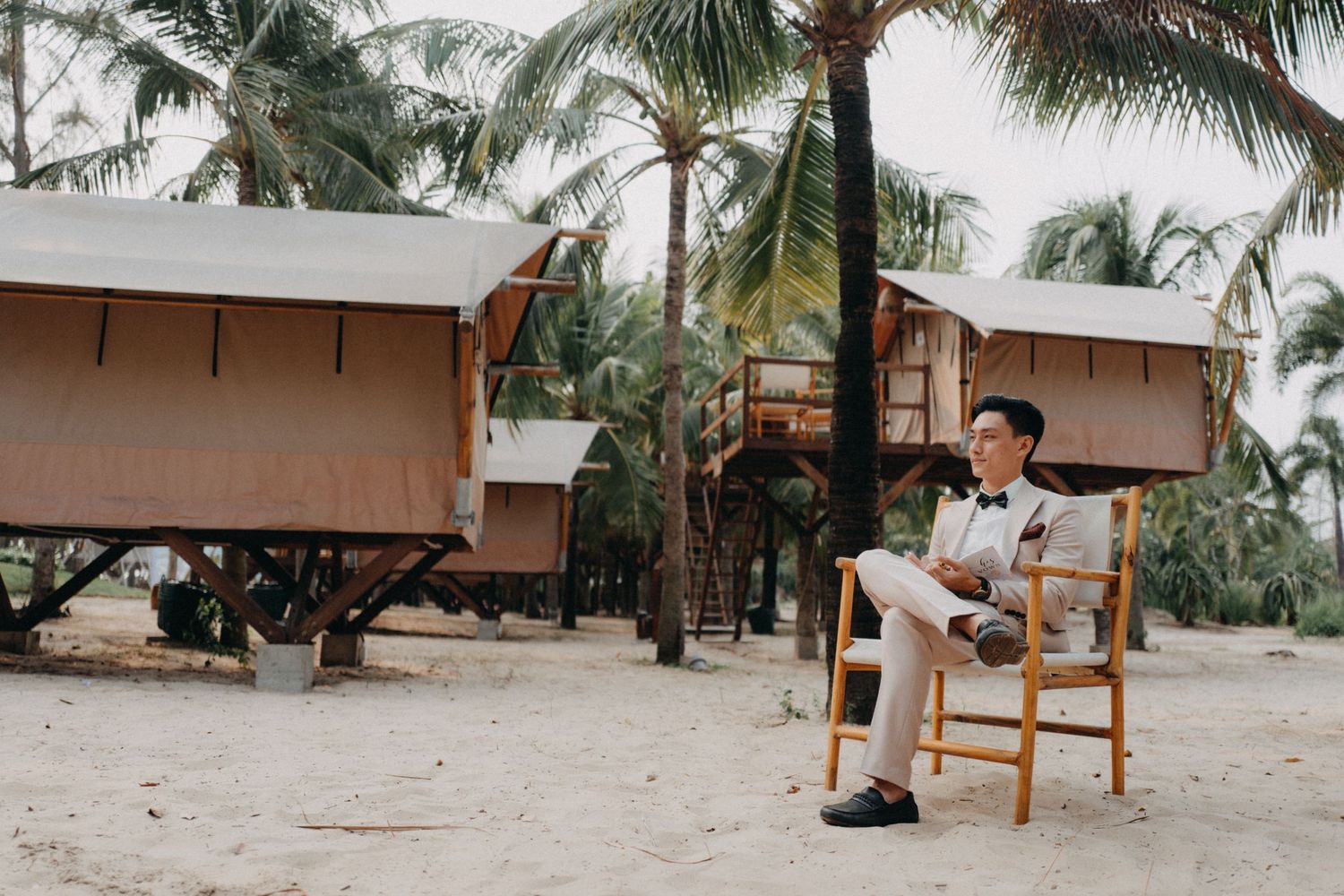 Groom in light suit sitting on chair near beach bungalows surrounded by tropical palm trees.