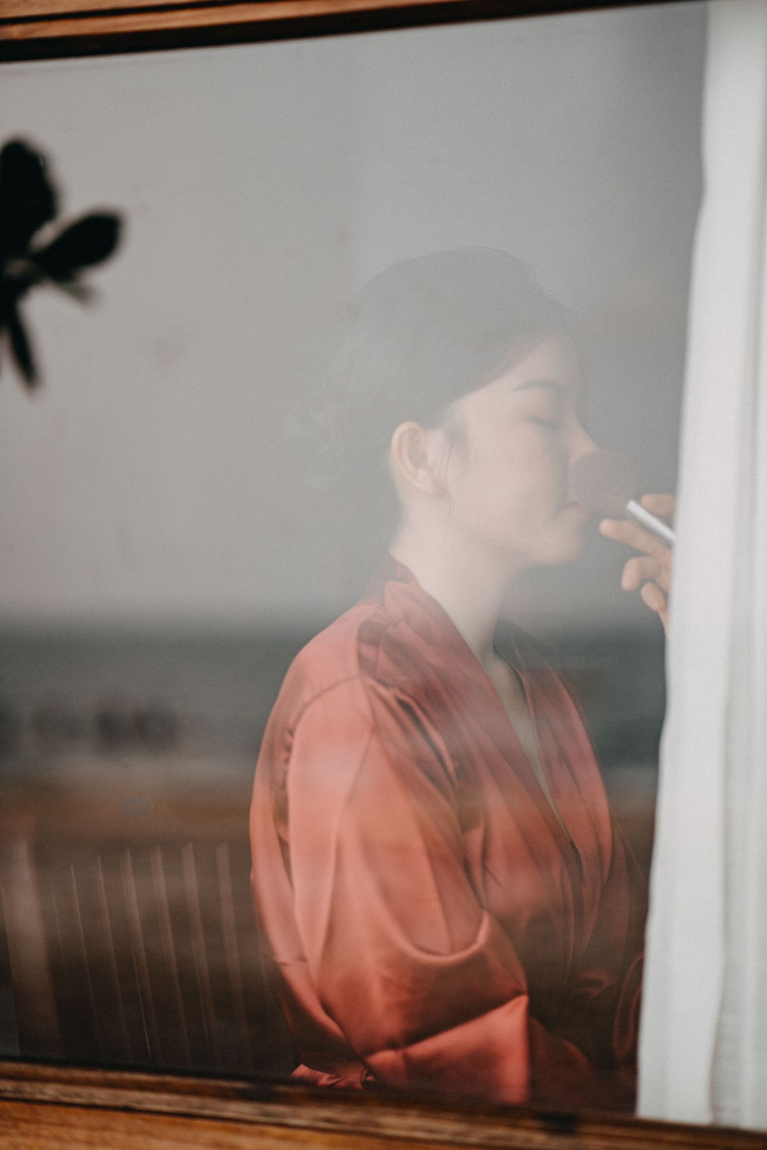 Bride in pink robe seen through misty window with ocean view in soft morning light.