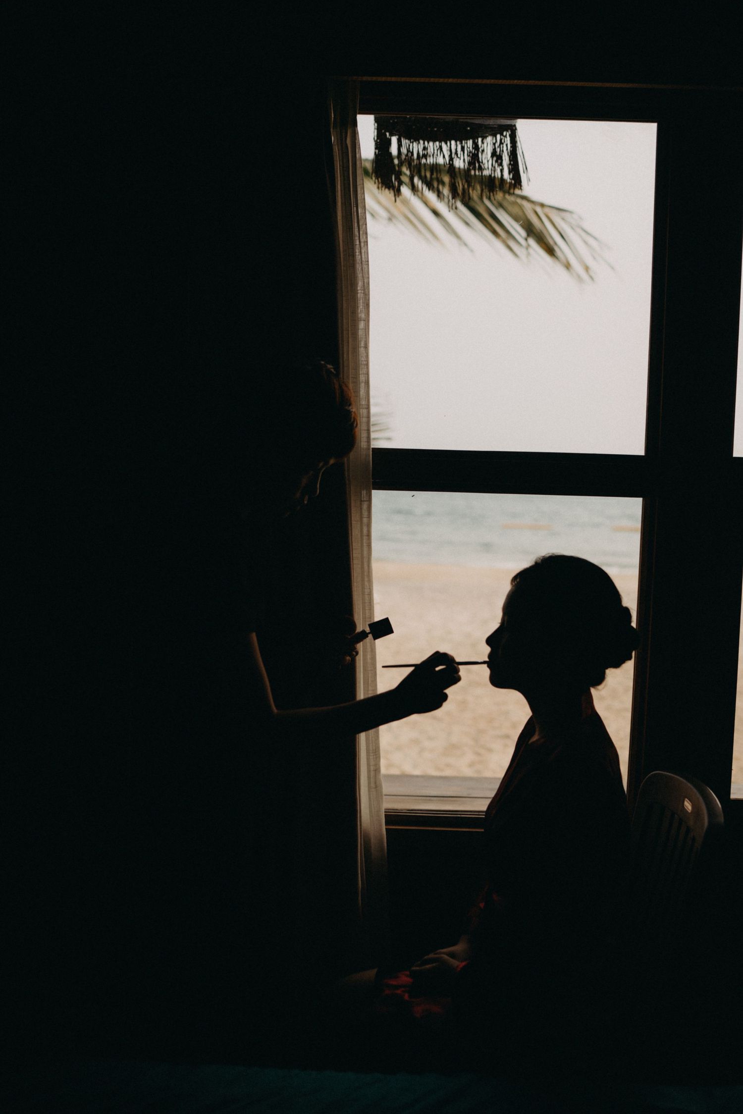Silhouette of bride getting makeup done by window with tropical beach view.