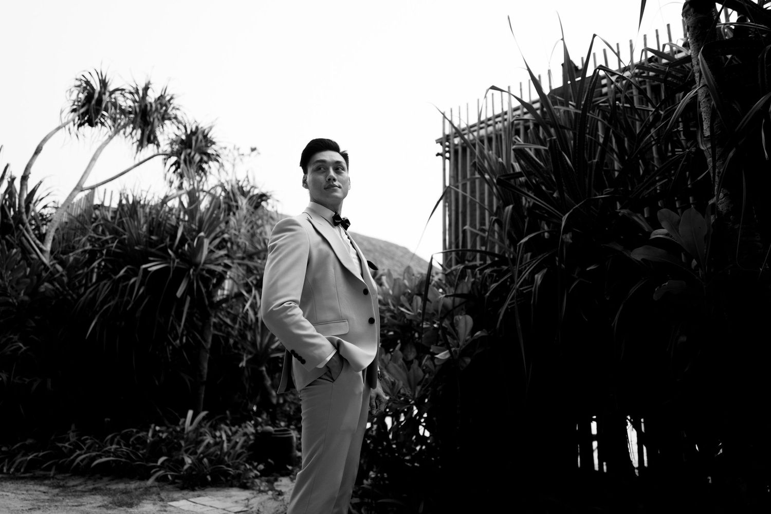 Elegant groom in gray suit posing among tropical plants in black and white.