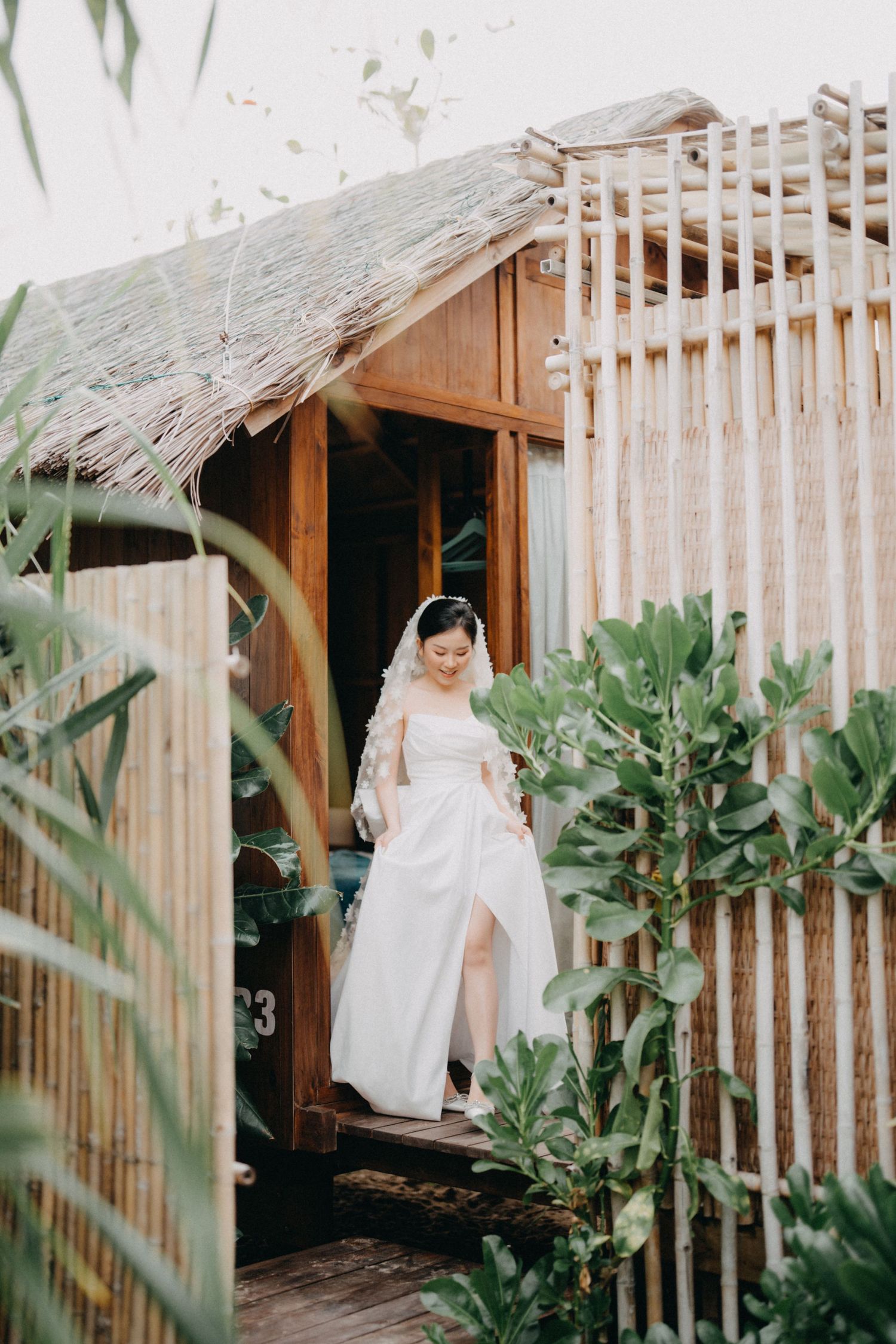 Bride in white gown and veil stepping out of bamboo tropical villa.