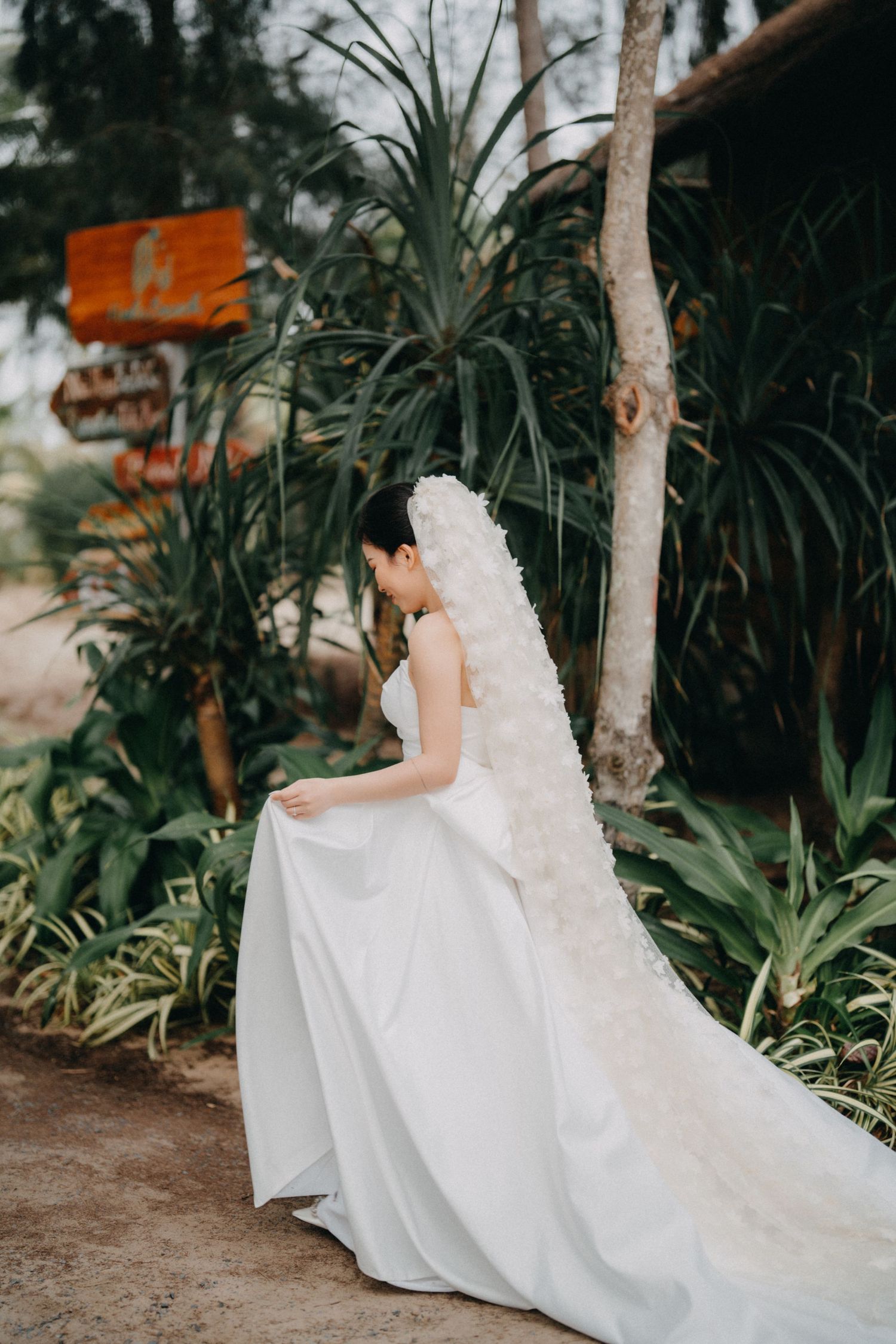 Bride in a white gown and floral veil poses gracefully in a lush tropical garden setting.