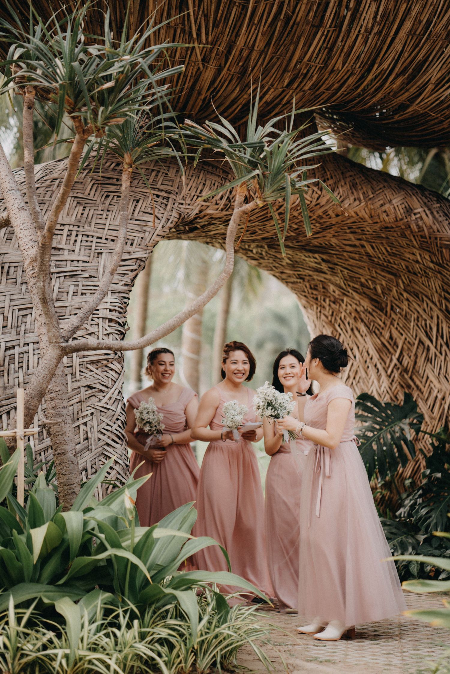 Four bridesmaids in blush pink dresses laugh together under a woven bamboo arch with tropical plants.