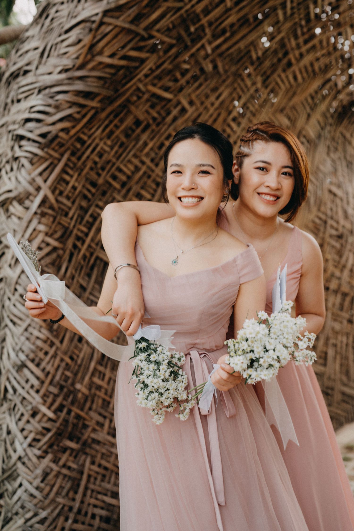 Two smiling bridesmaids in pink dresses hold white bouquets against a rustic woven bamboo backdrop.