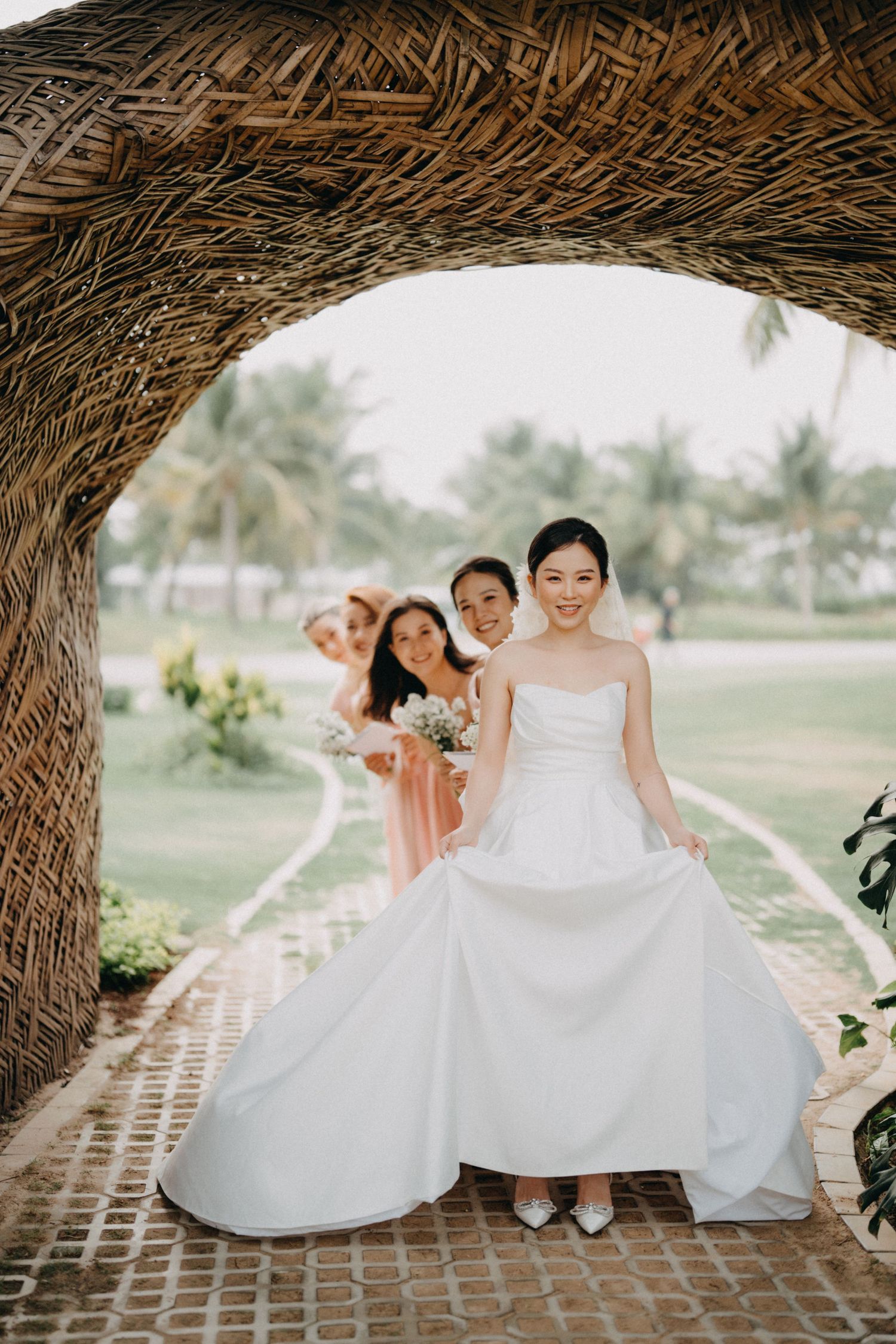 Bride in white strapless gown leads bridesmaids through a woven bamboo archway at a tropical venue.
