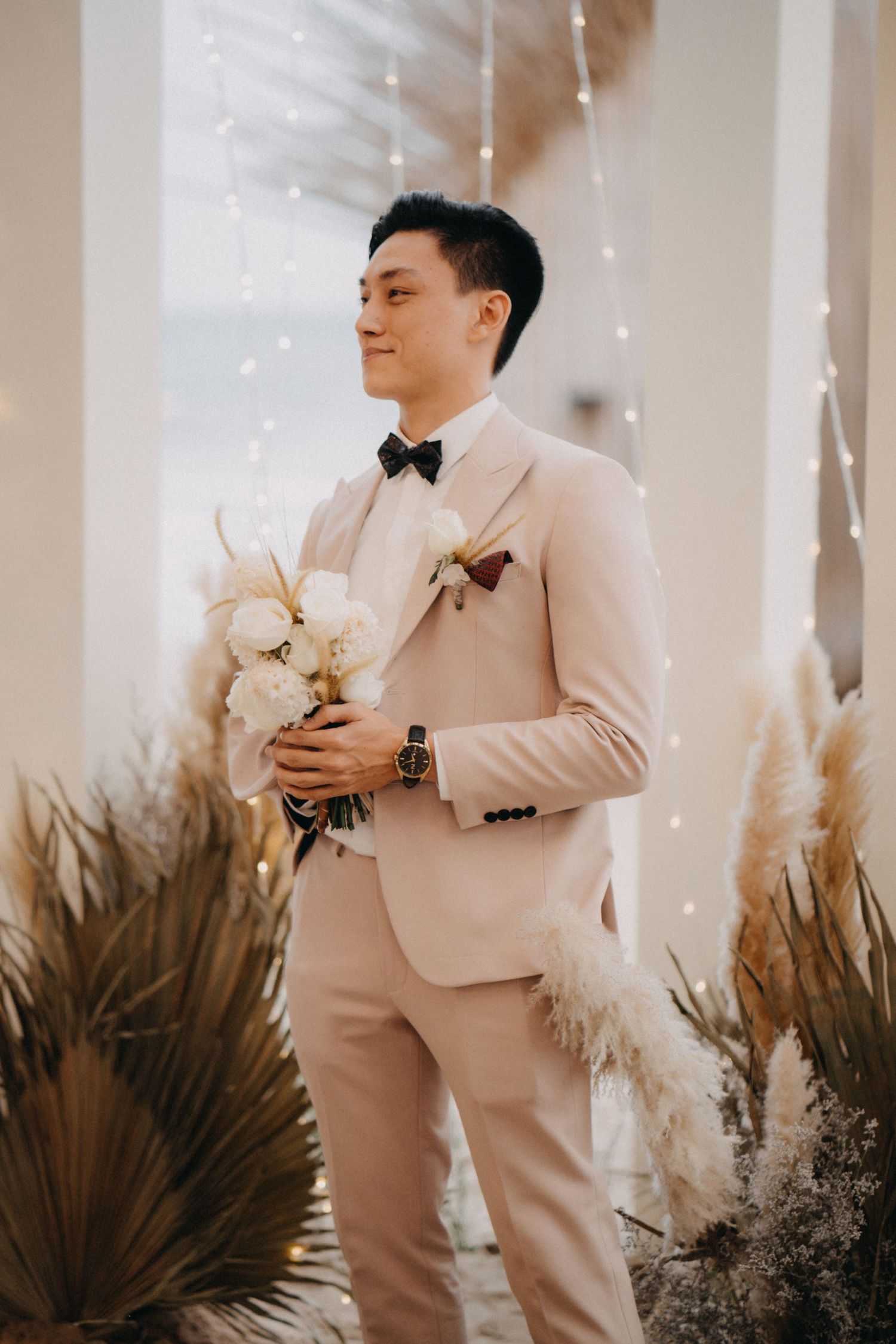 Dapper groom in a beige suit holds a white bouquet surrounded by pampas grass and fairy lights.