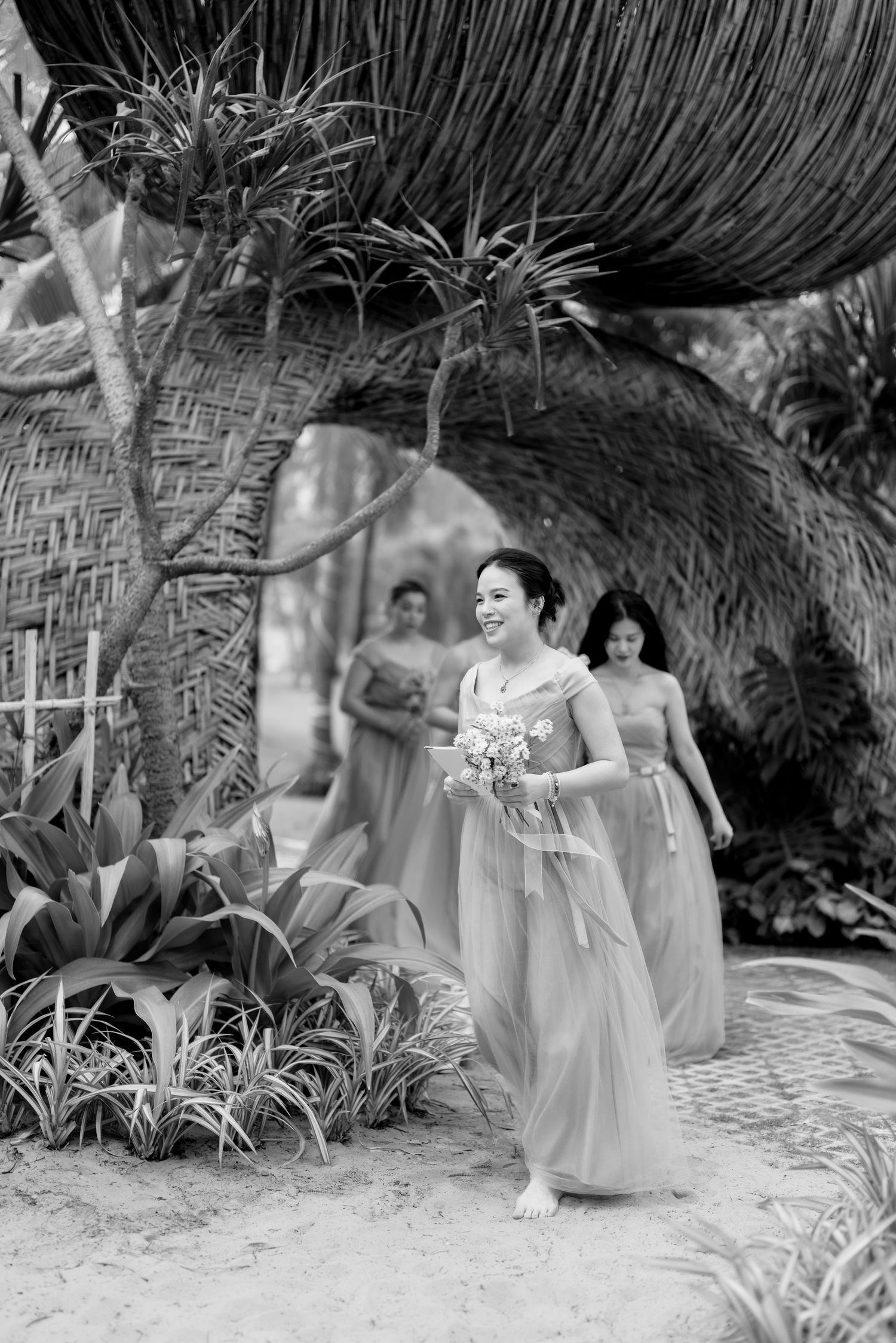 Bridesmaids in flowing pink dresses walk through a tropical bamboo archway in black and white photo.