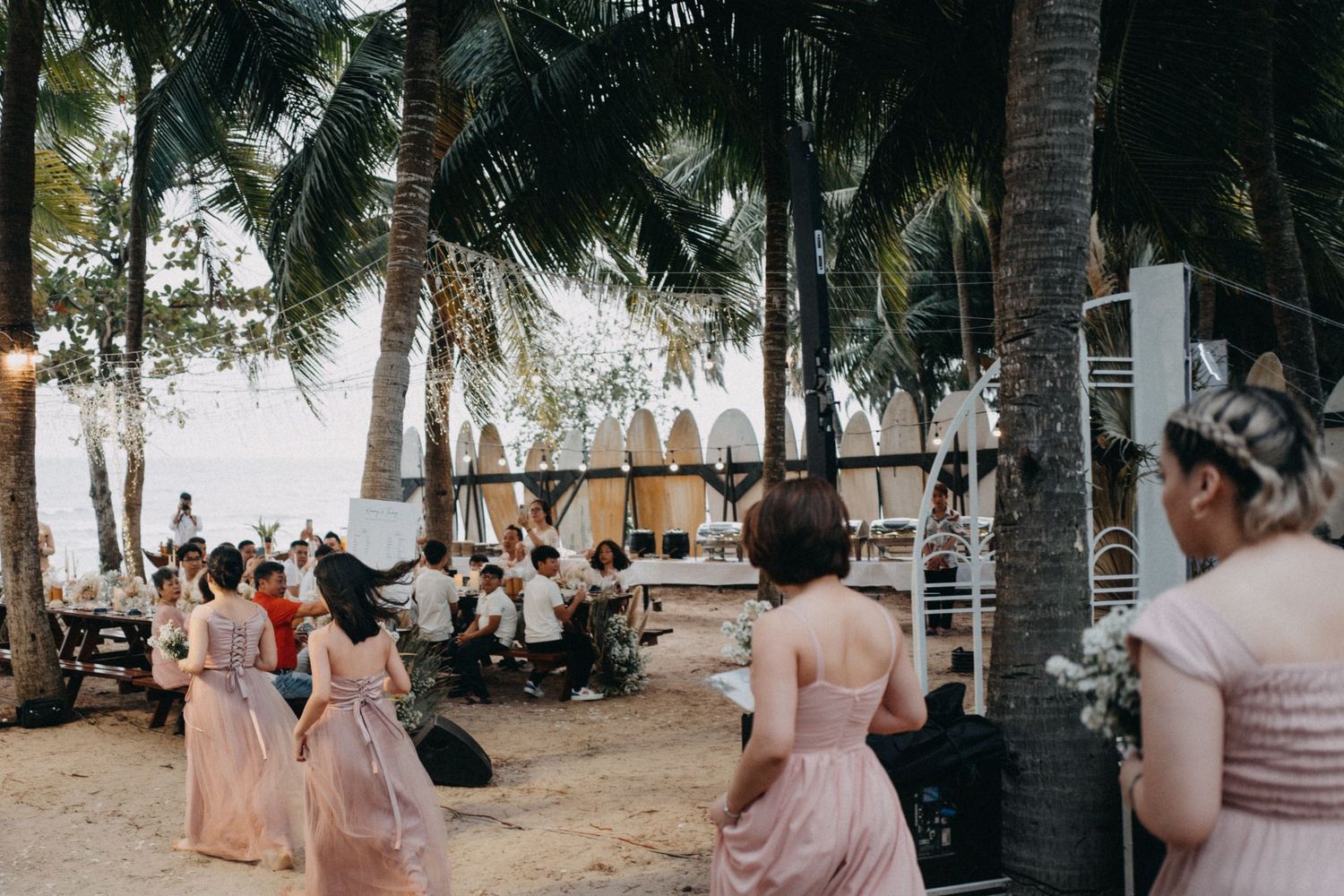 Bridesmaids in blush dresses gather near palm trees at a beachside wedding ceremony setup.